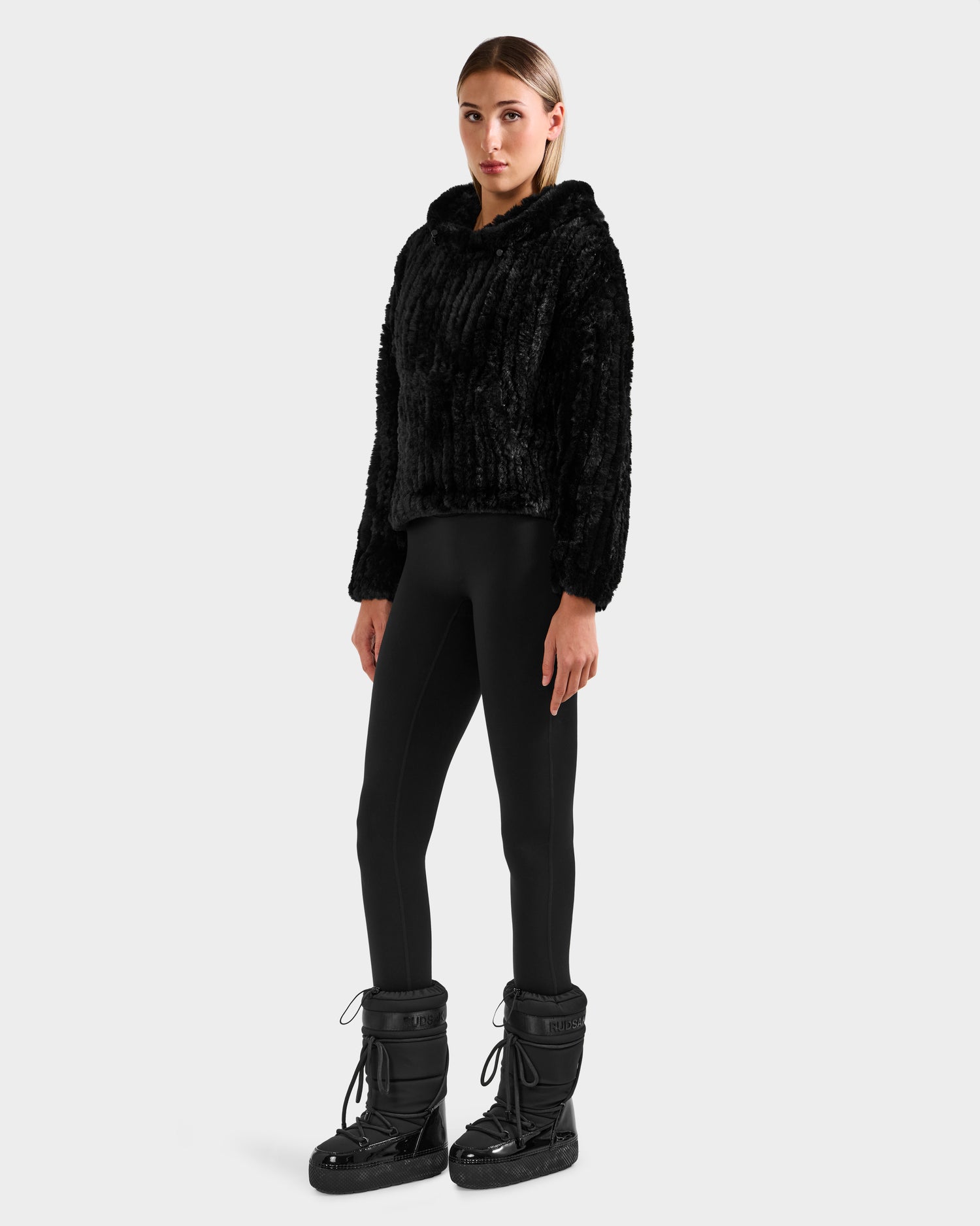 A woman faces forward, wearing the Rudsak AUGUST - 8525658 Black faux fur hoodie—ideal for winter lounging—paired with black leggings and padded boots, set against a plain light background.