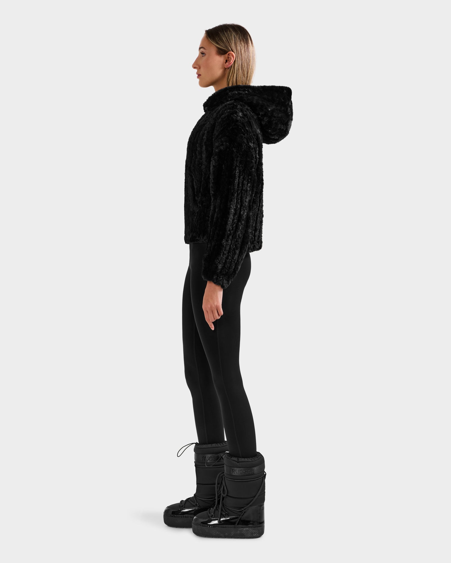 A woman stands in profile wearing the Rudsak AUGUST - 8525658 Black faux fur hoodie, styled with black leggings and winter boots, set against a plain light gray background—ideal for cozy winter lounging.