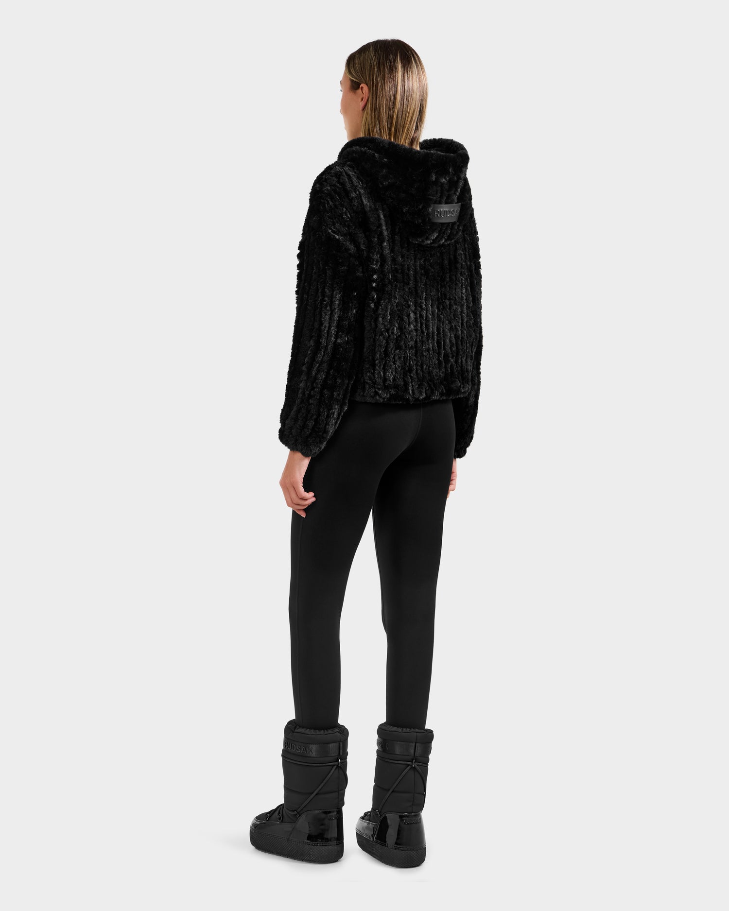 A woman faces away, wearing the Rudsak AUGUST - 8525658 Black textured faux fur hoodie jacket, black leggings, and winter boots against a light gray background—ideal for après-ski or cozy winter lounging.