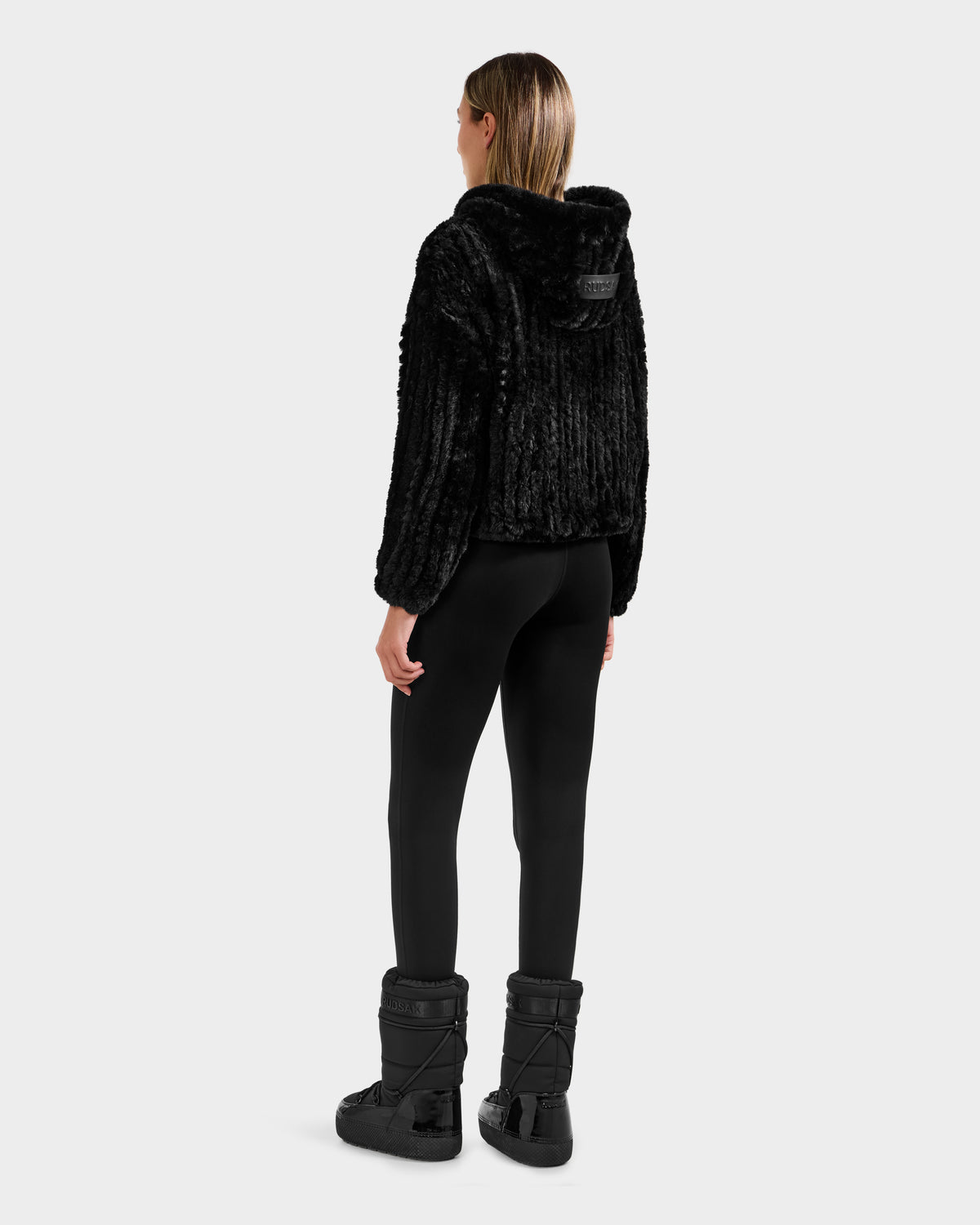 A woman faces away, wearing the Rudsak AUGUST - 8525658 Black textured faux fur hoodie jacket, black leggings, and winter boots against a light gray background—ideal for après-ski or cozy winter lounging.