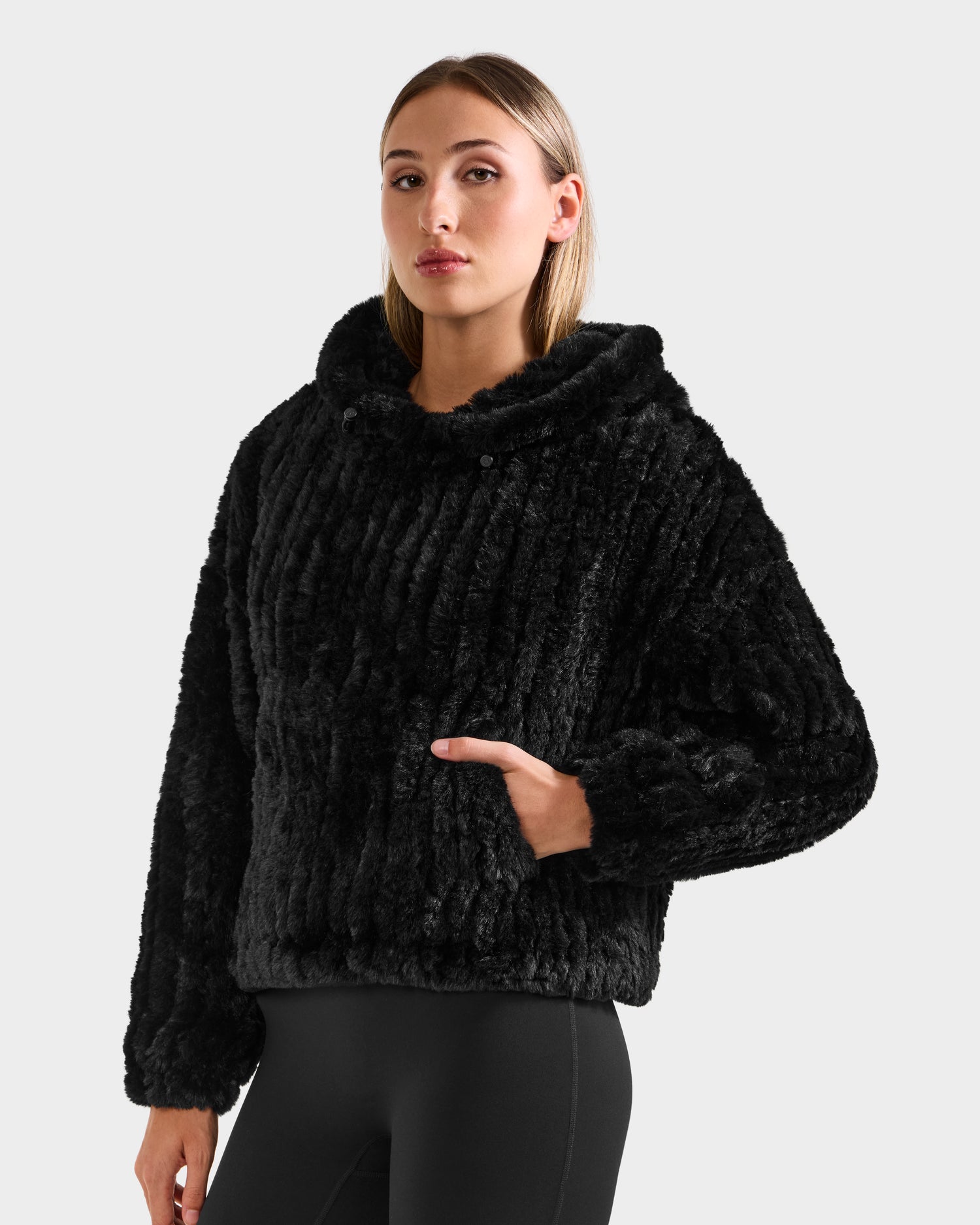 A person wears the Rudsak AUGUST - 8525658 Black faux fur hoodie with black leggings, standing against a light gray background—ideal for winter lounging or cozy après-ski moments.