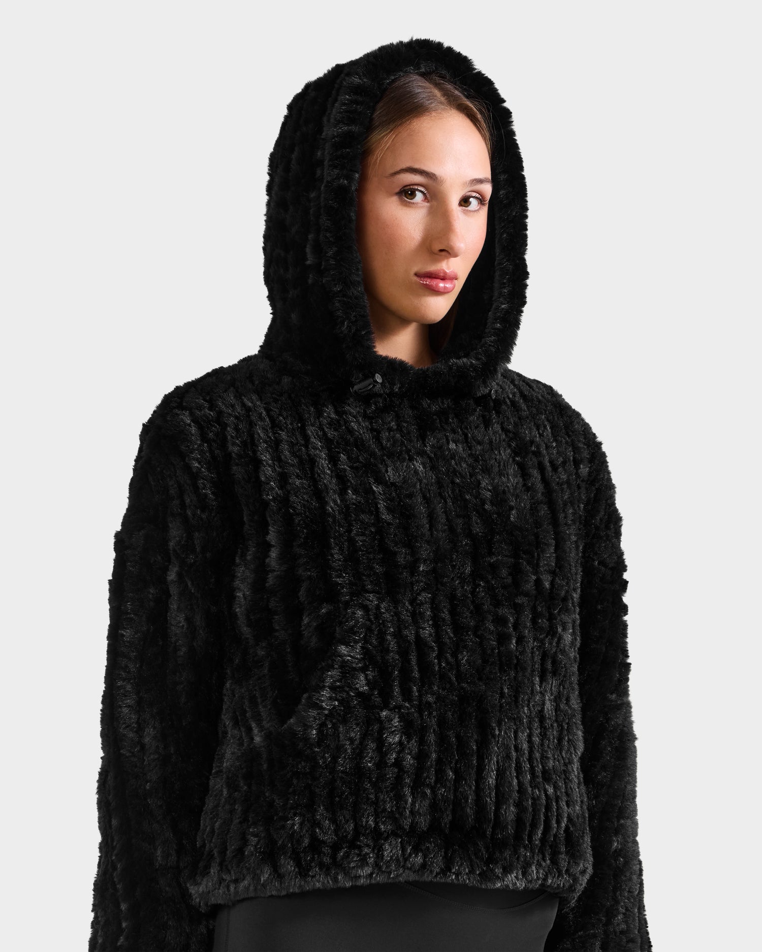A woman models the Rudsak AUGUST - 8525658 Black textured faux fur hoodie against a light gray background, gazing to the side with a neutral expression—ideal for winter lounging or chic après-ski style.