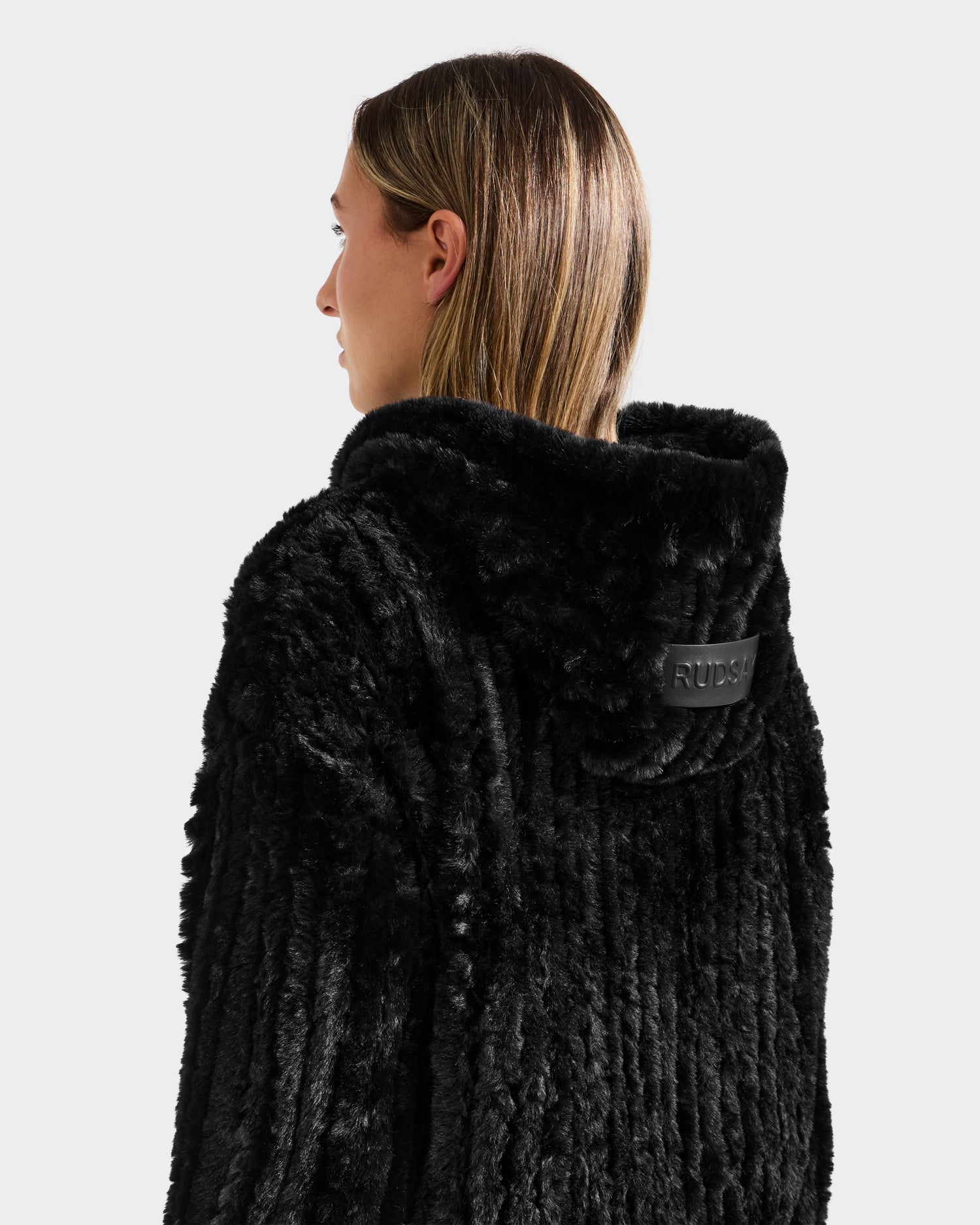A person with straight, shoulder-length hair is seen from behind wearing the Rudsak AUGUST - 8525658 Black faux fur hoodie, featuring a hood and black patch—ideal for winter lounging or après-ski style.