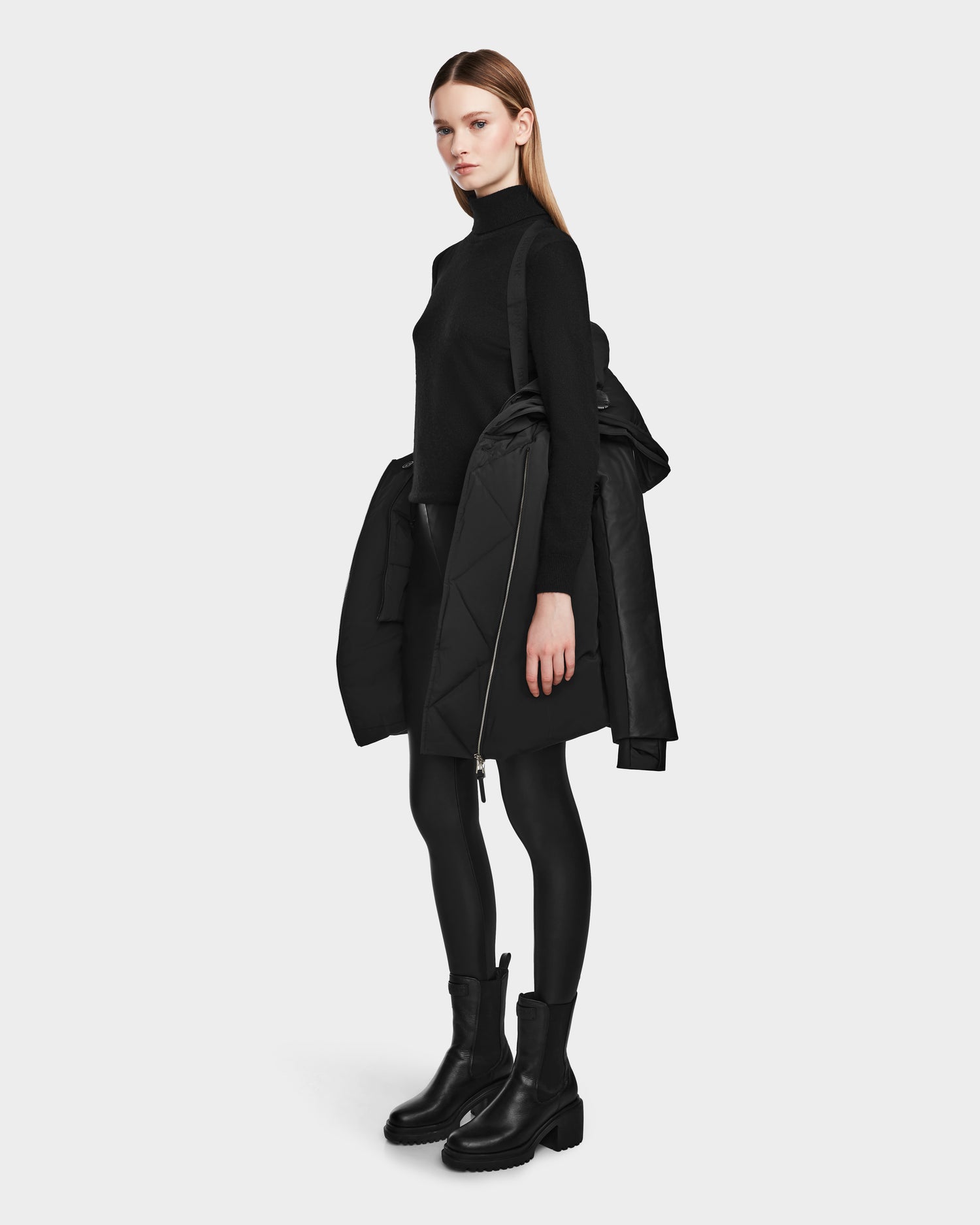 A woman stands sideways against a white background, holding the Rudsak AURELLE - L124538 Black women’s down parka with vegan leather sleeves over her arm while wearing a black turtleneck, skirt, leggings, and boots.