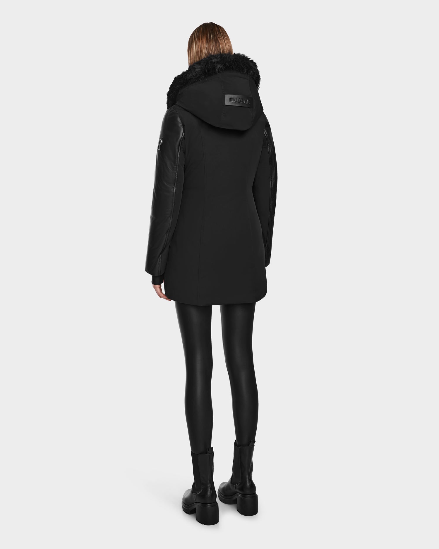 A person faces away, wearing the Rudsak AURELLE - L124538 Black women’s down parka with vegan leather sleeves and fur-trimmed hood, paired with black leggings and boots—offering stylish winter protection against a light background.