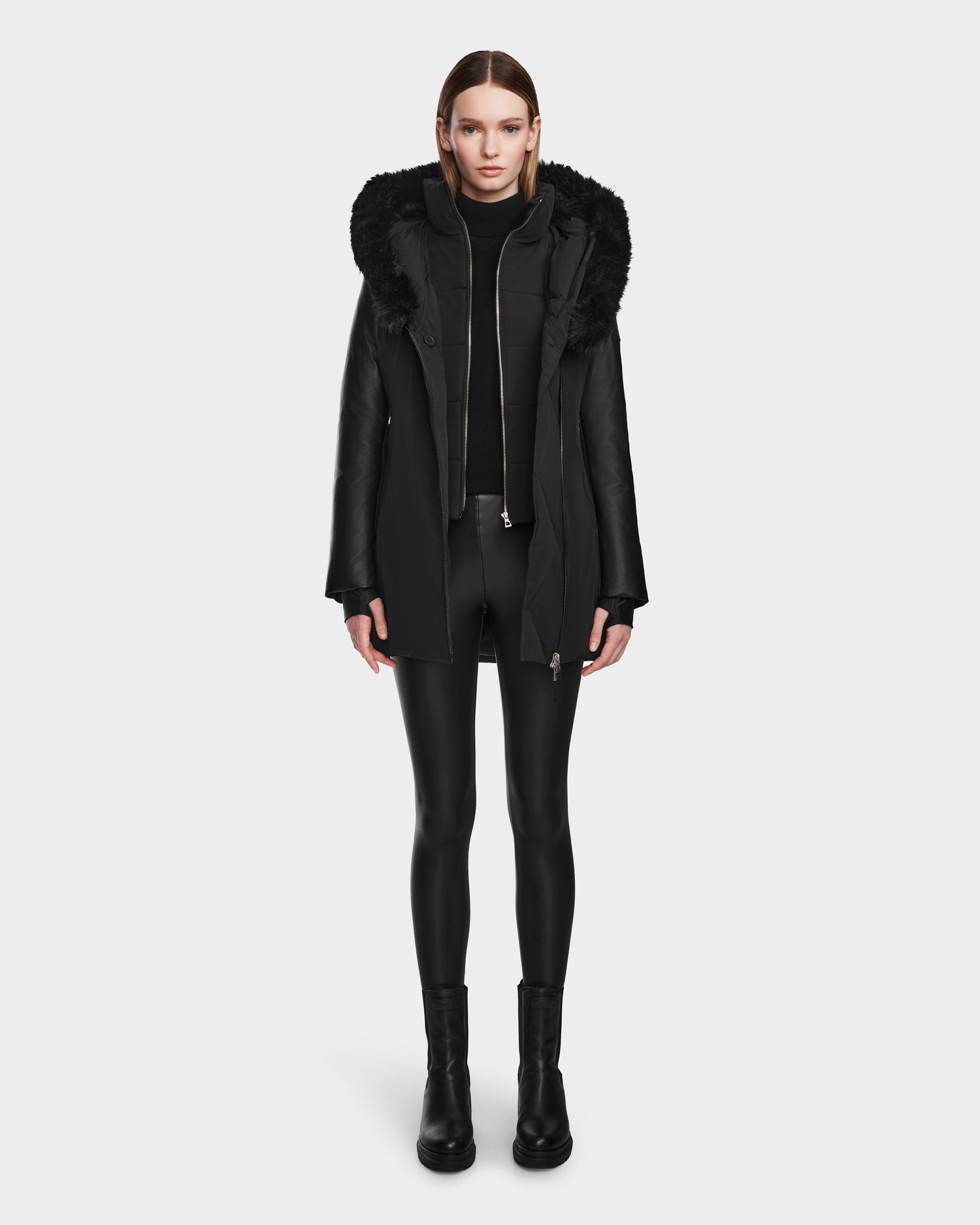 A woman faces forward wearing the Rudsak AURELLE - L124538 Black womens down parka with vegan leather sleeves and a fur-lined hood, styled with a black turtleneck, leggings, and boots against a plain white background.