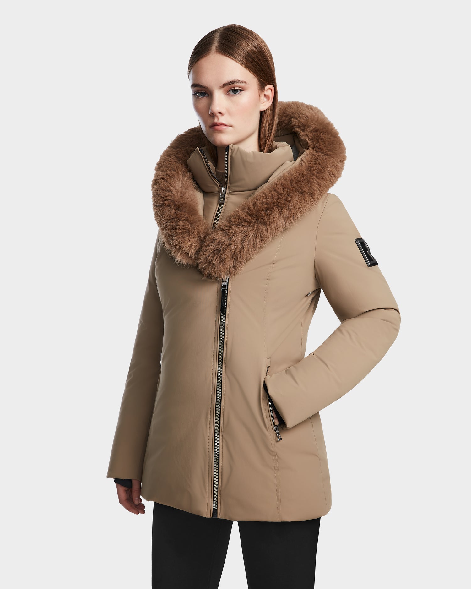 A woman models the Rudsak AURORE - L123561 Sand, a tan fitted winter coat with a large brown faux fur-trimmed hood, standing against a plain white background.