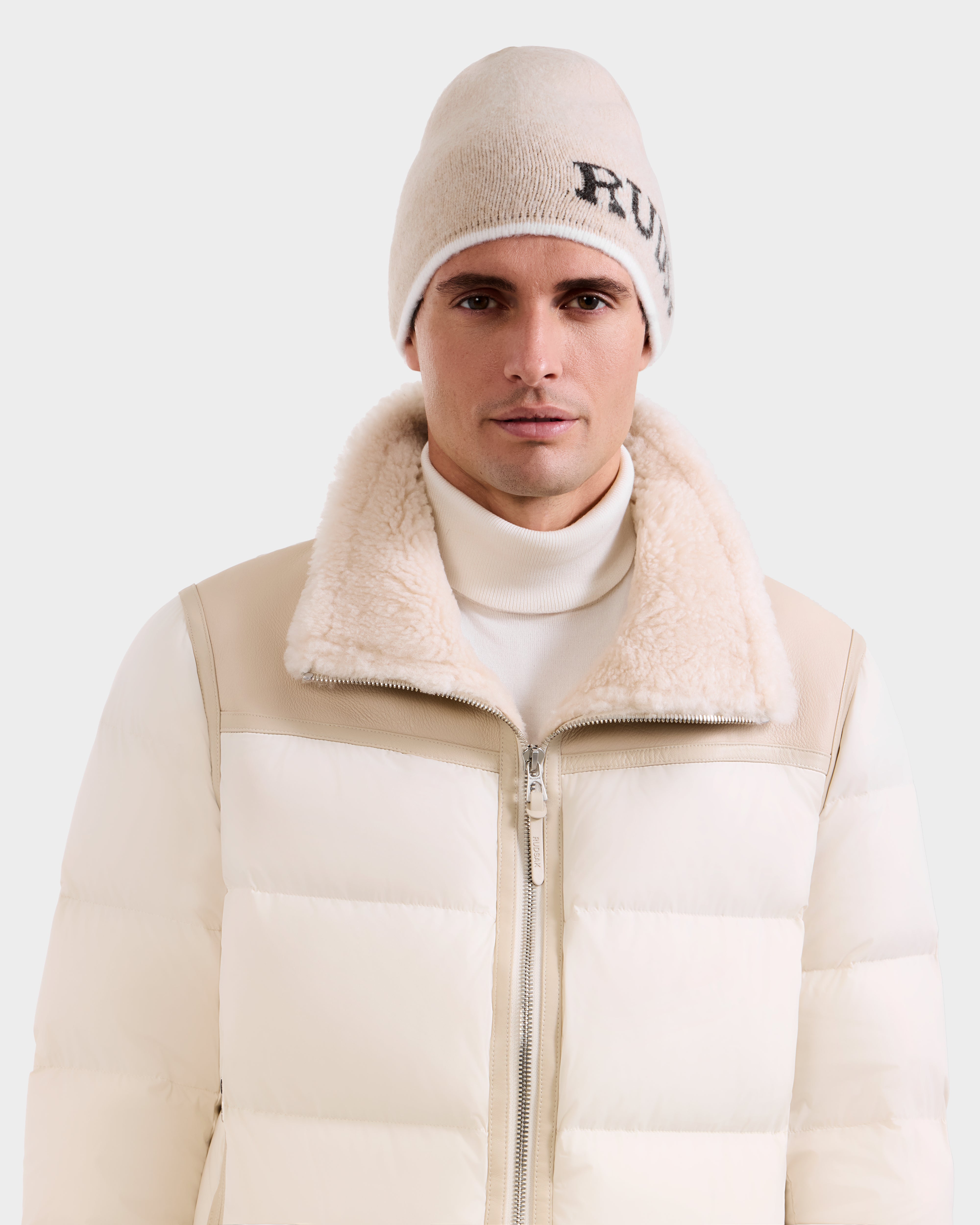 A man wears the Rudsak AXEL - 6425040 Vanilla men’s beanie with black text, a shearling-collared puffer jacket, and a white turtleneck sweater, layered in cozy winter style against a plain light background.