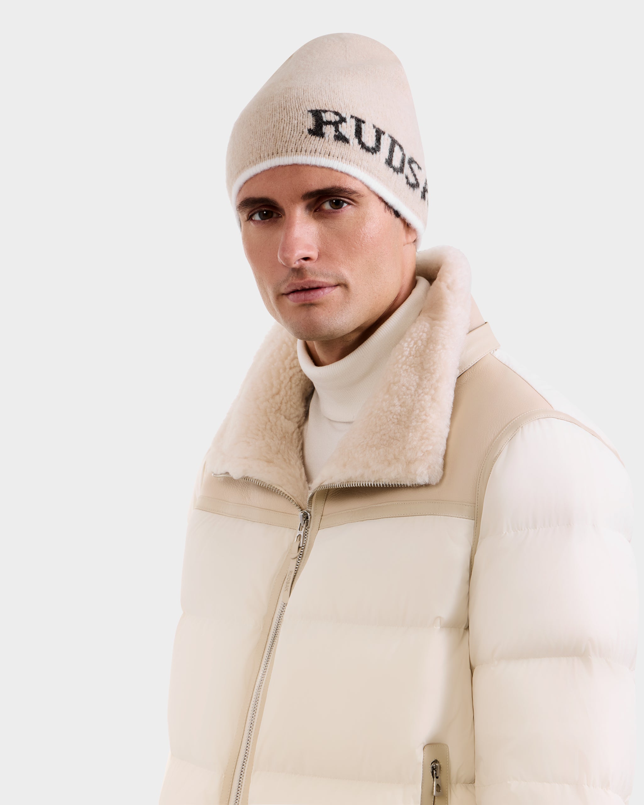A man wears a Rudsak AXEL - 6425040 Vanilla cream puffer jacket with shearling collar over a white turtleneck, paired with a light beige Rudsak men’s beanie, posing against a white background—ideal winter style.