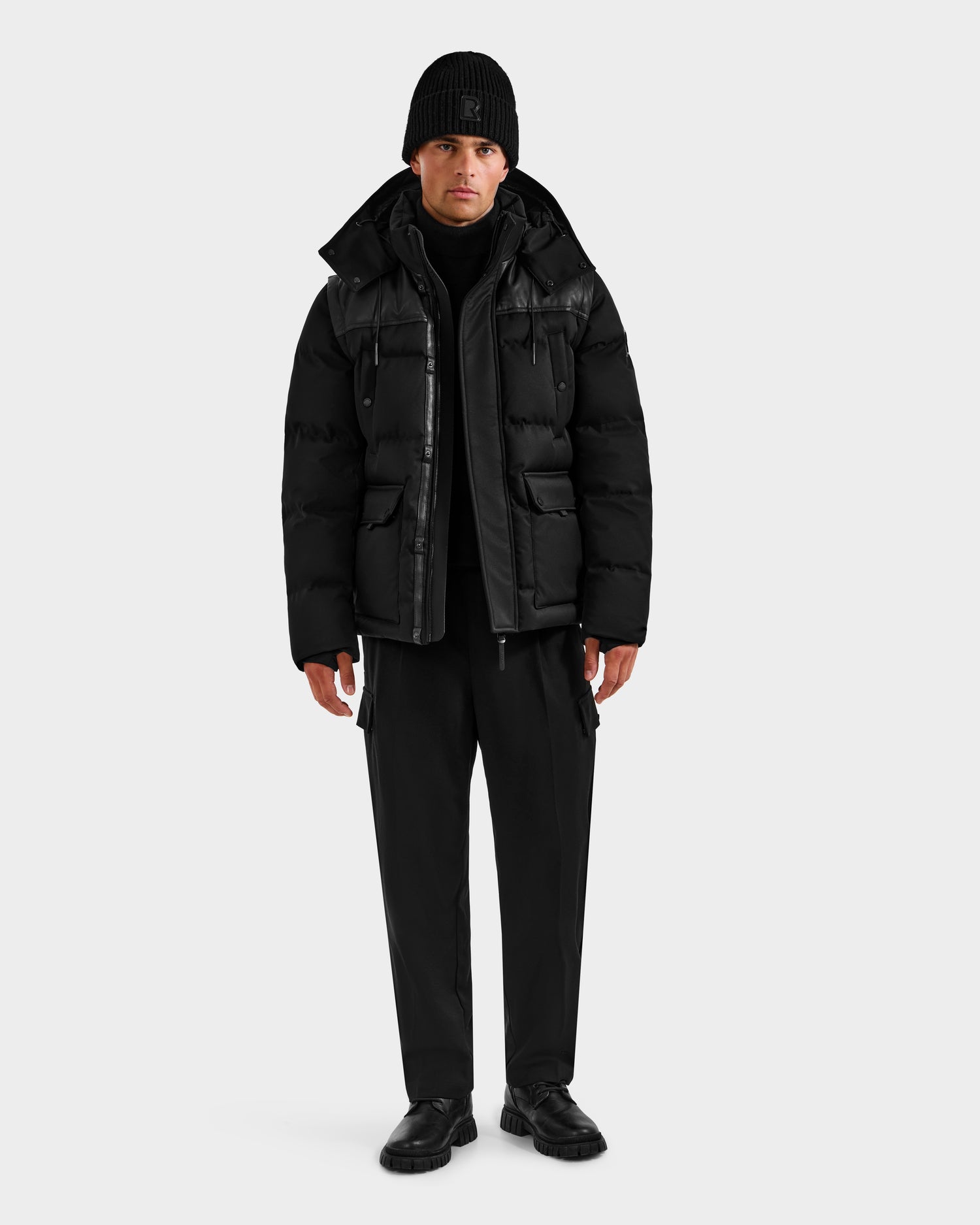 A man stands facing forward in a Rudsak BALDWIN - 6124507 Black RDS-certified down men’s parka with multiple pockets, black beanie, pants, and shoes, against a plain light background.