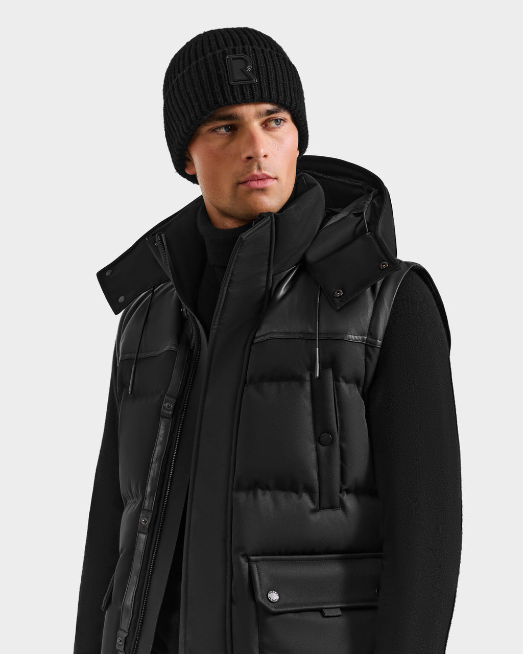 A man wearing the Rudsak BALDWIN - 6124507 Black puffer vest with removable sleeves and a high collar, paired with a black beanie and sweater, looks to the side against a plain light gray background.