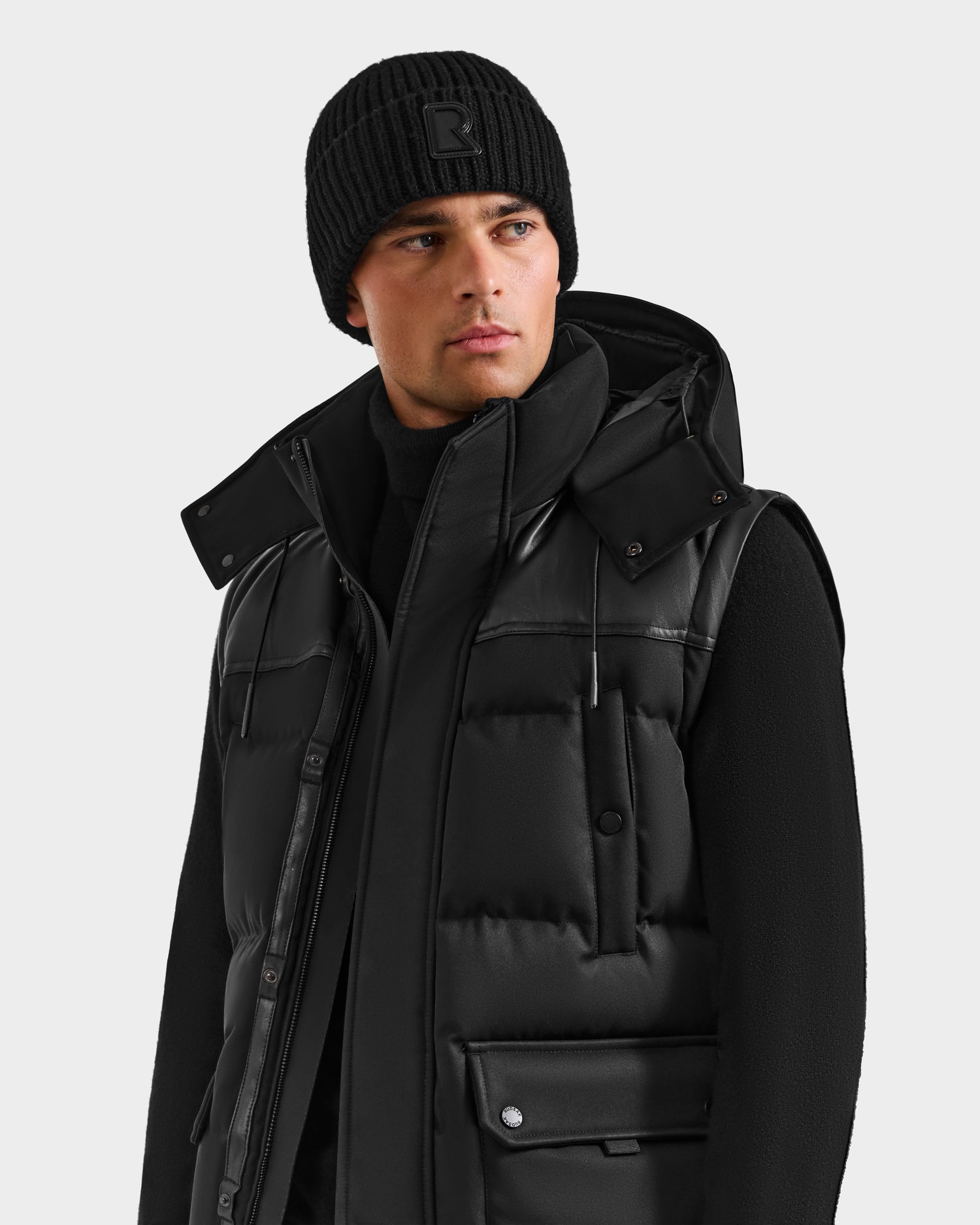 A man wearing the Rudsak BALDWIN - 6124507 Black puffer vest with removable sleeves and a high collar, paired with a black beanie and sweater, looks to the side against a plain light gray background.