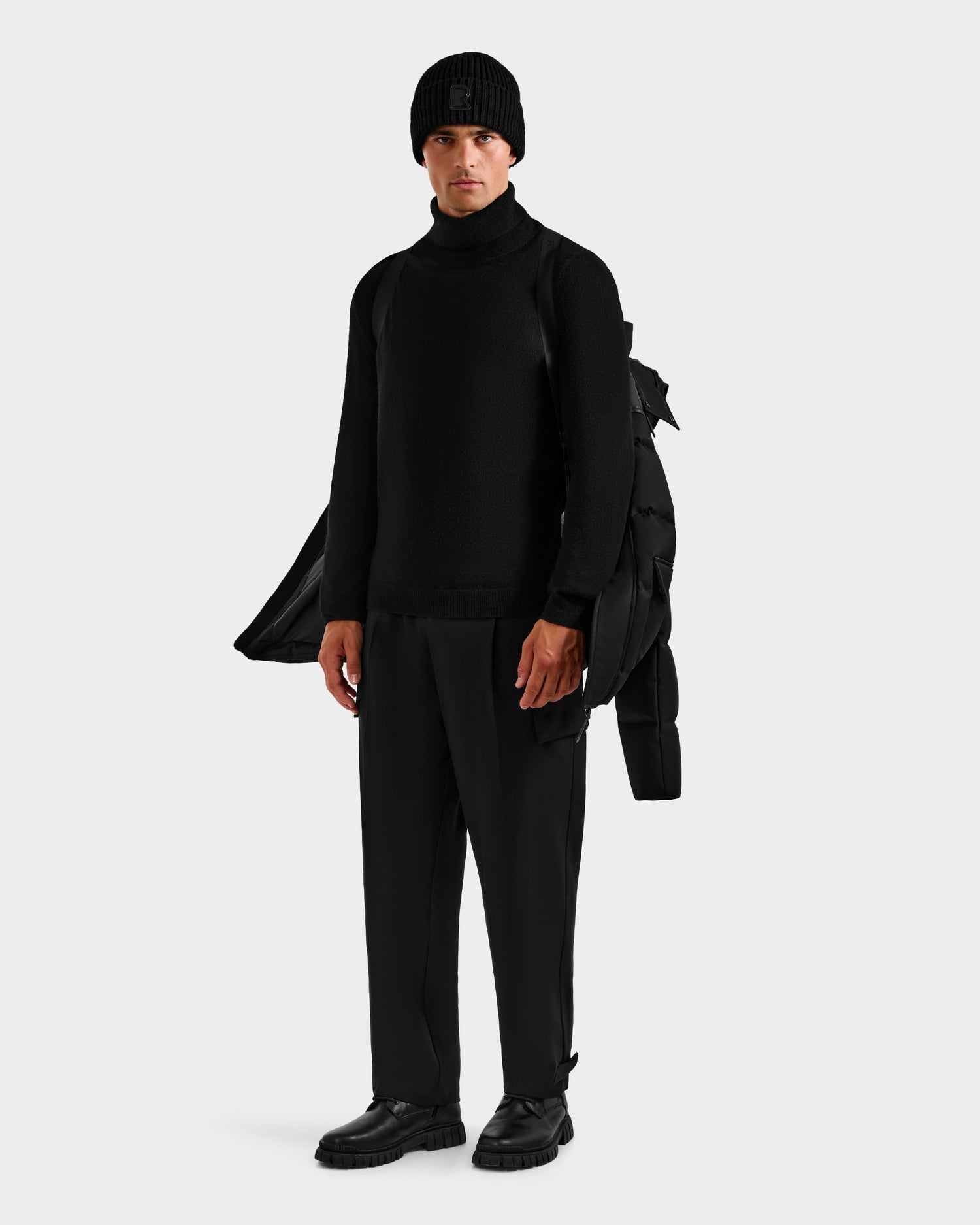 A person stands against a plain background wearing black clothing and holding the Rudsak BALDWIN - 6124507 Black men's down parka with removable sleeves over one shoulder.