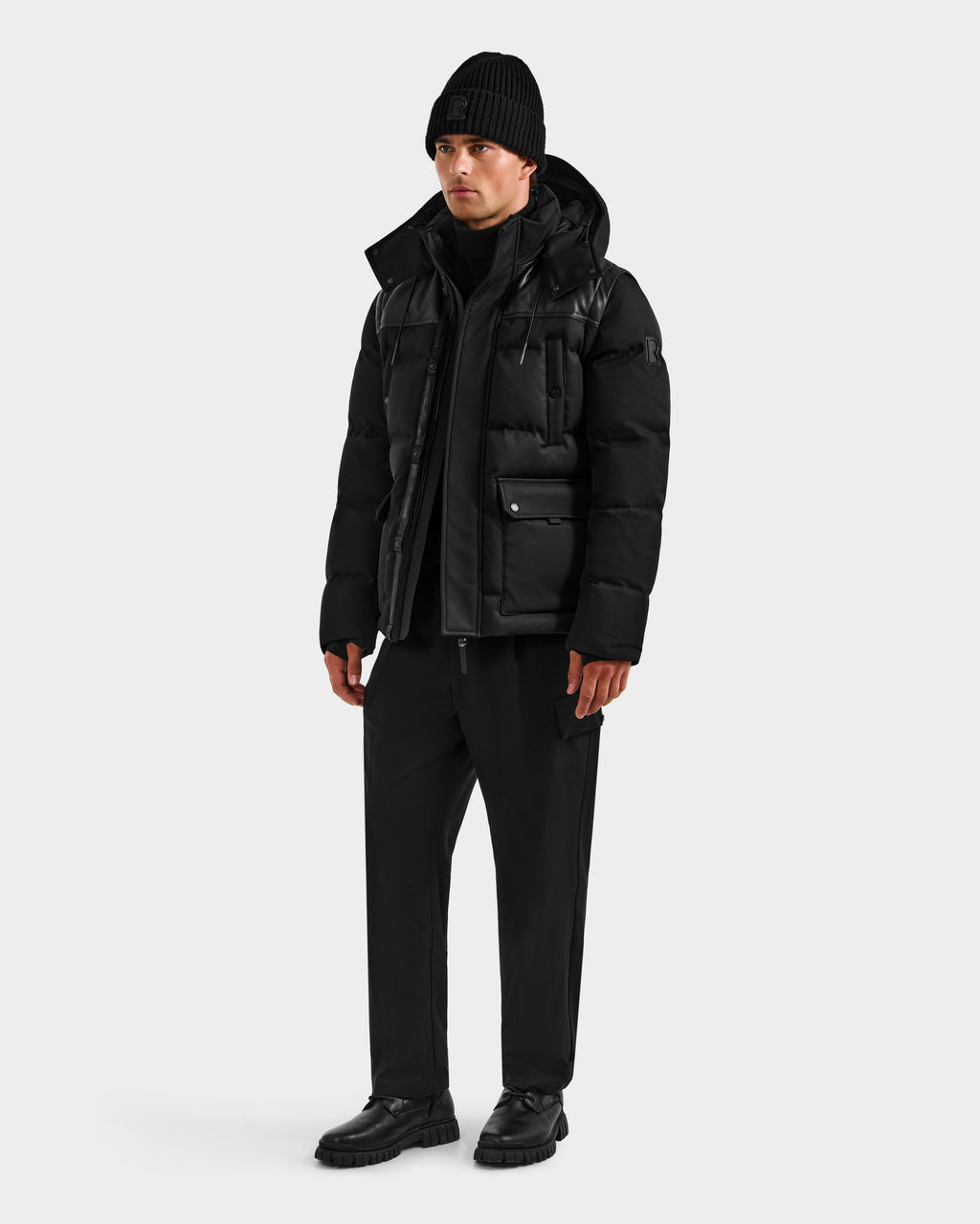 A person stands against a plain background wearing the BALDWIN - 6124507 Black men’s down parka by Rudsak—styled with a black beanie, layers, pants, and shoes for an all-black, winter-ready look.