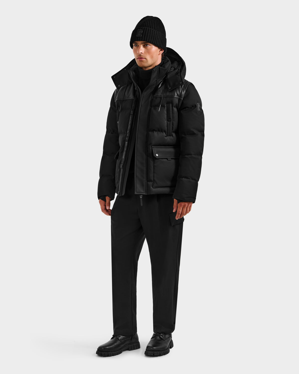 A person stands against a plain background wearing the BALDWIN - 6124507 Black men’s down parka by Rudsak—styled with a black beanie, layers, pants, and shoes for an all-black, winter-ready look.