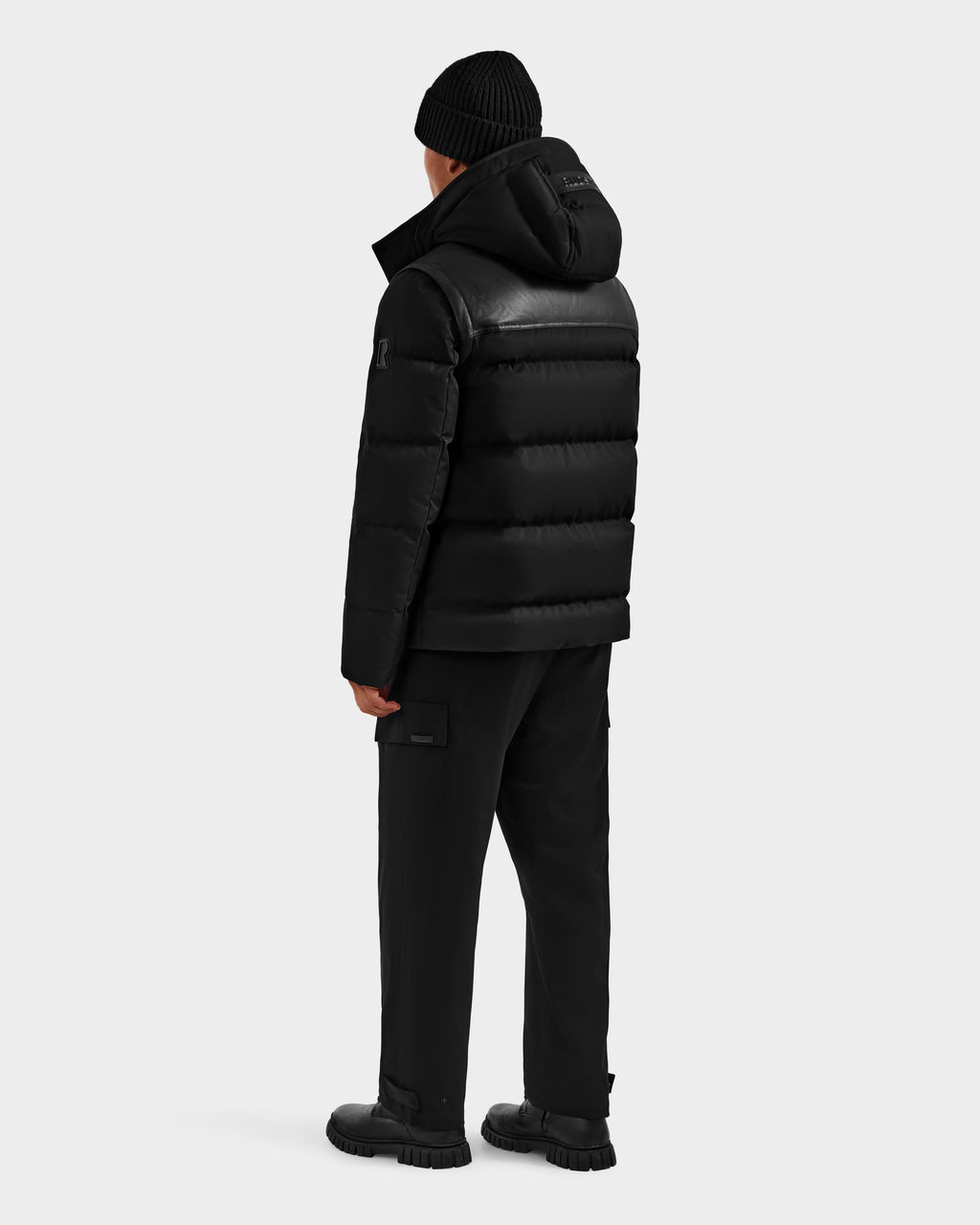 A person faces away, wearing the Rudsak BALDWIN - 6124507 Black men's down parka with removable sleeves, styled with black cargo pants, boots, and a black beanie against a plain light background.