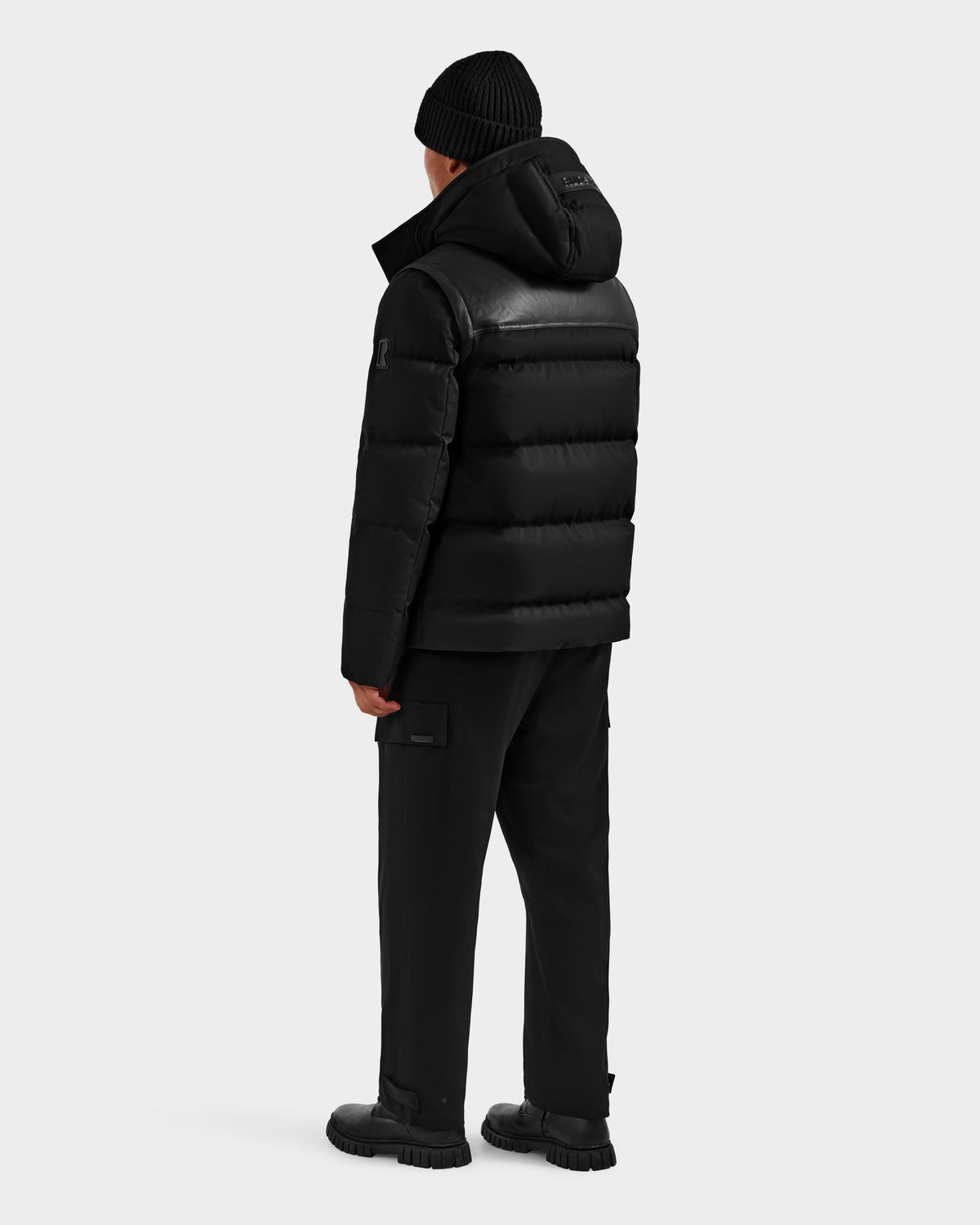 A person faces away, wearing the Rudsak BALDWIN - 6124507 Black men's down parka with removable sleeves, styled with black cargo pants, boots, and a black beanie against a plain light background.