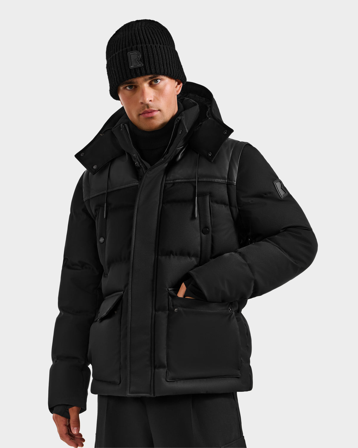 A man wears the Rudsak BALDWIN - 6124507 Black men’s down parka with large pockets and a matching black beanie, standing against a plain light background.