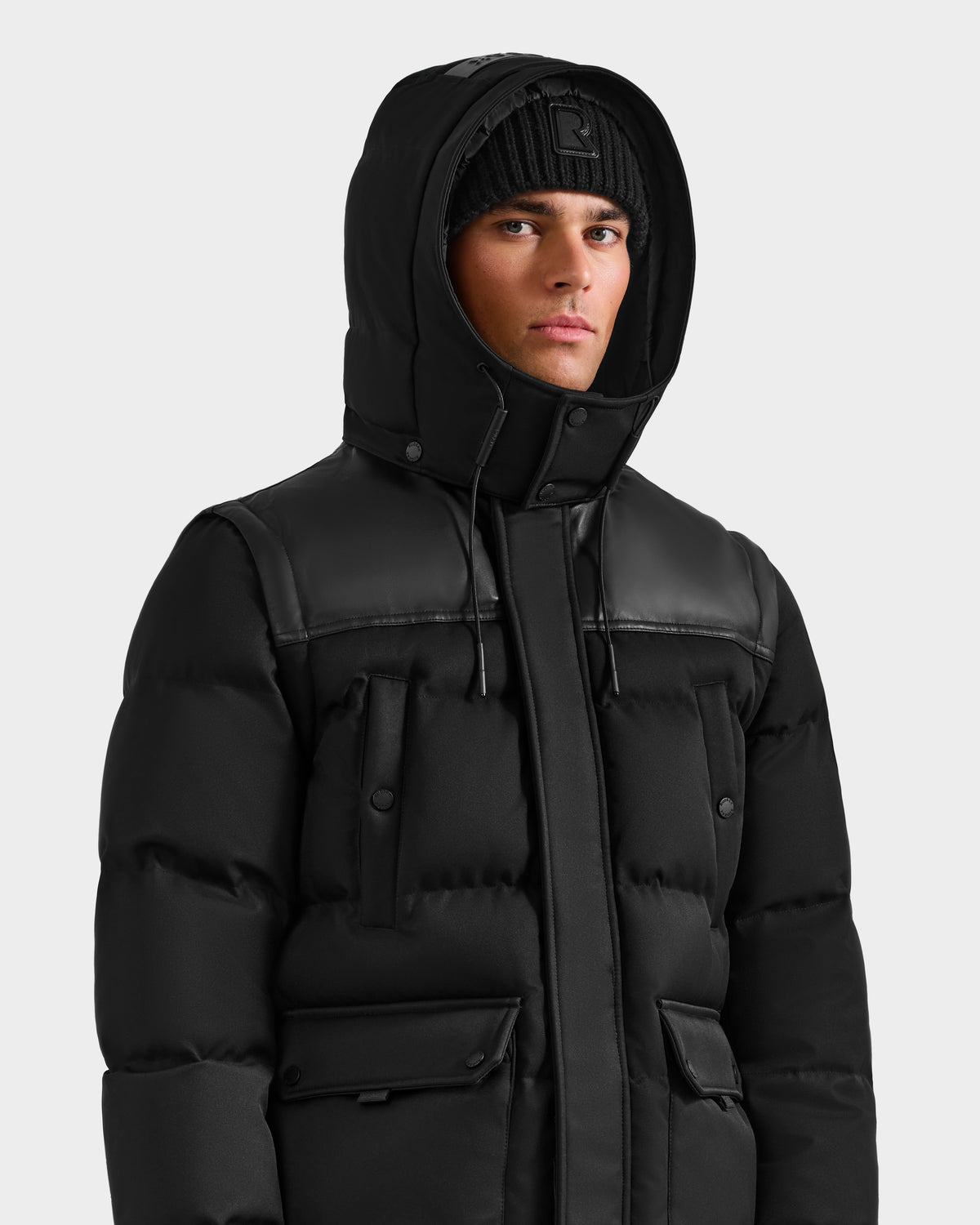 A person wears the Rudsak BALDWIN - 6124507 Black men’s down parka with buttoned pockets and removable sleeves, paired with a black knit beanie, facing forward against a plain light gray background.