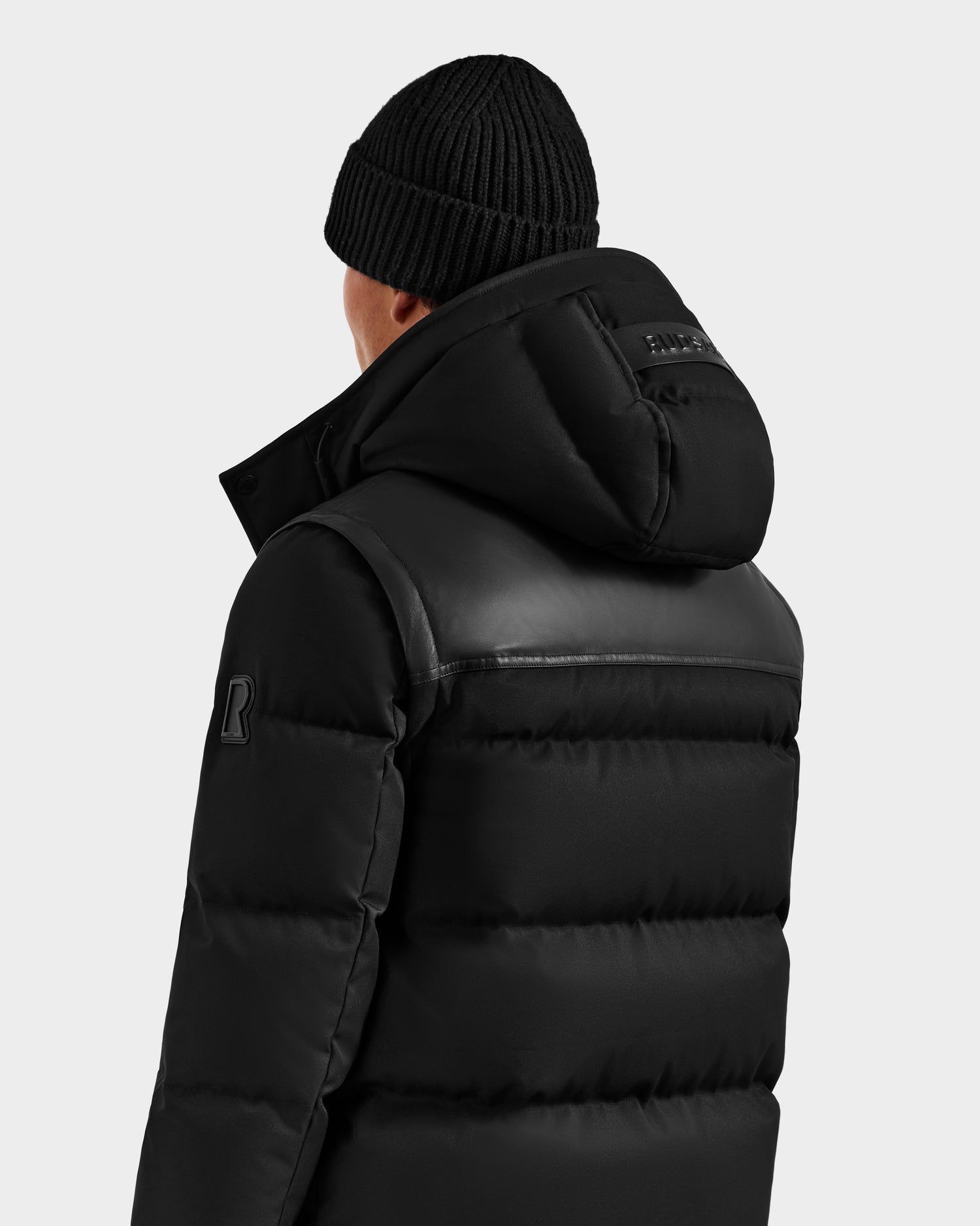 A person wearing the Rudsak BALDWIN - 6124507 black men’s down parka with hood and a black knit beanie, shown from the back against a plain light background.