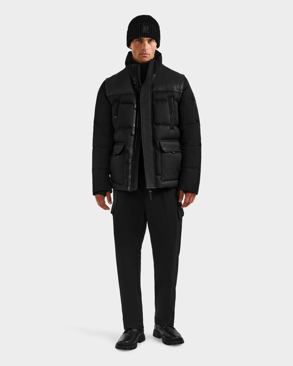 A man stands facing forward in the Rudsak BALDWIN - 6124507 Black men’s down parka with removable sleeves, paired with black pants and shoes, against a plain light gray background.