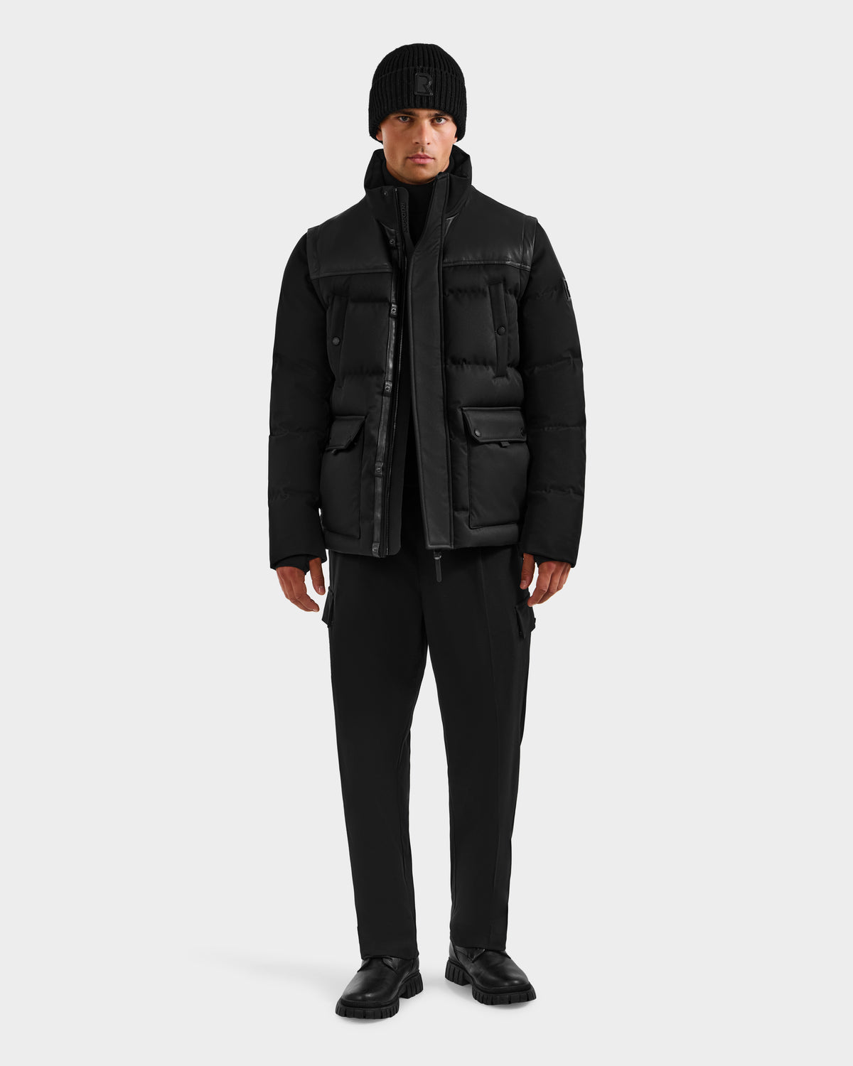 A man stands facing forward in the Rudsak BALDWIN - 6124507 Black men’s down parka with removable sleeves, paired with black pants and shoes, against a plain light gray background.