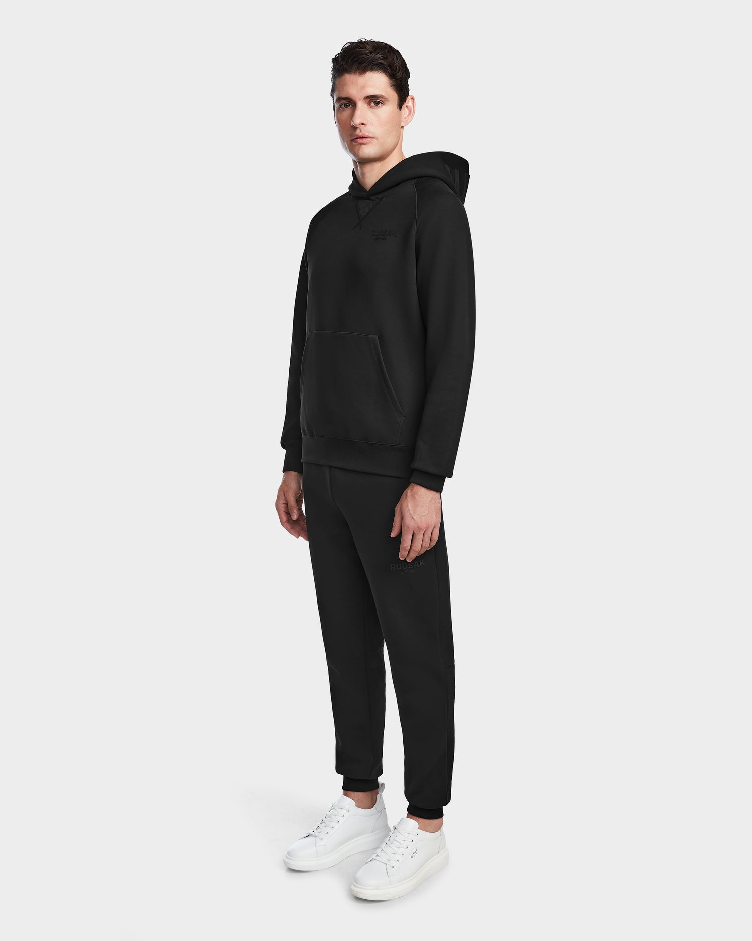 A person stands facing forward in a black Rudsak BARRY - 9524011 hoodie (size M) and black sweatpants with white sneakers, against a plain light background.