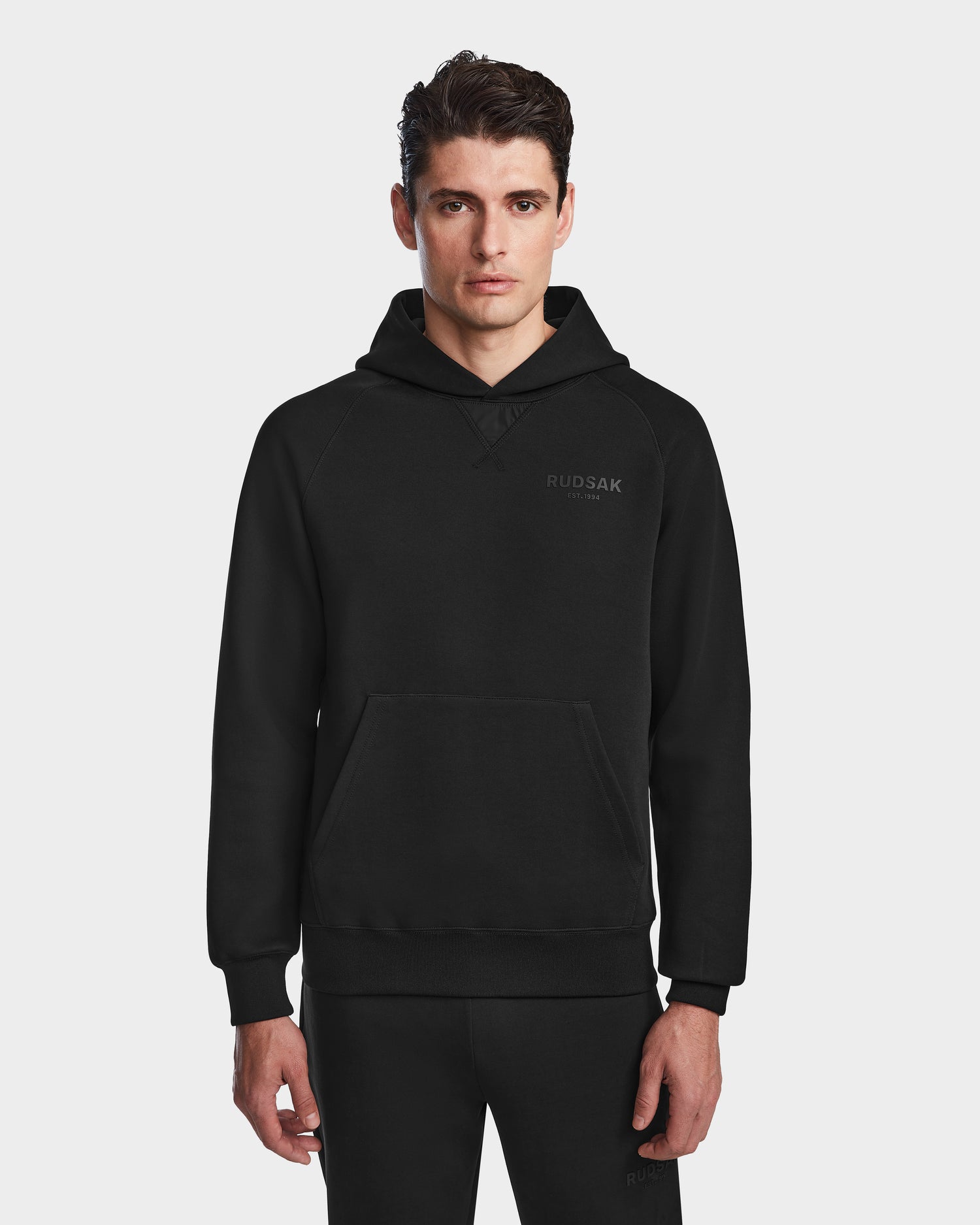 A man faces forward wearing Rudsak’s BARRY oversized unisex black hoodie (9524011, size M) with subtle chest branding and a front pocket, paired with matching pants. The background is white.