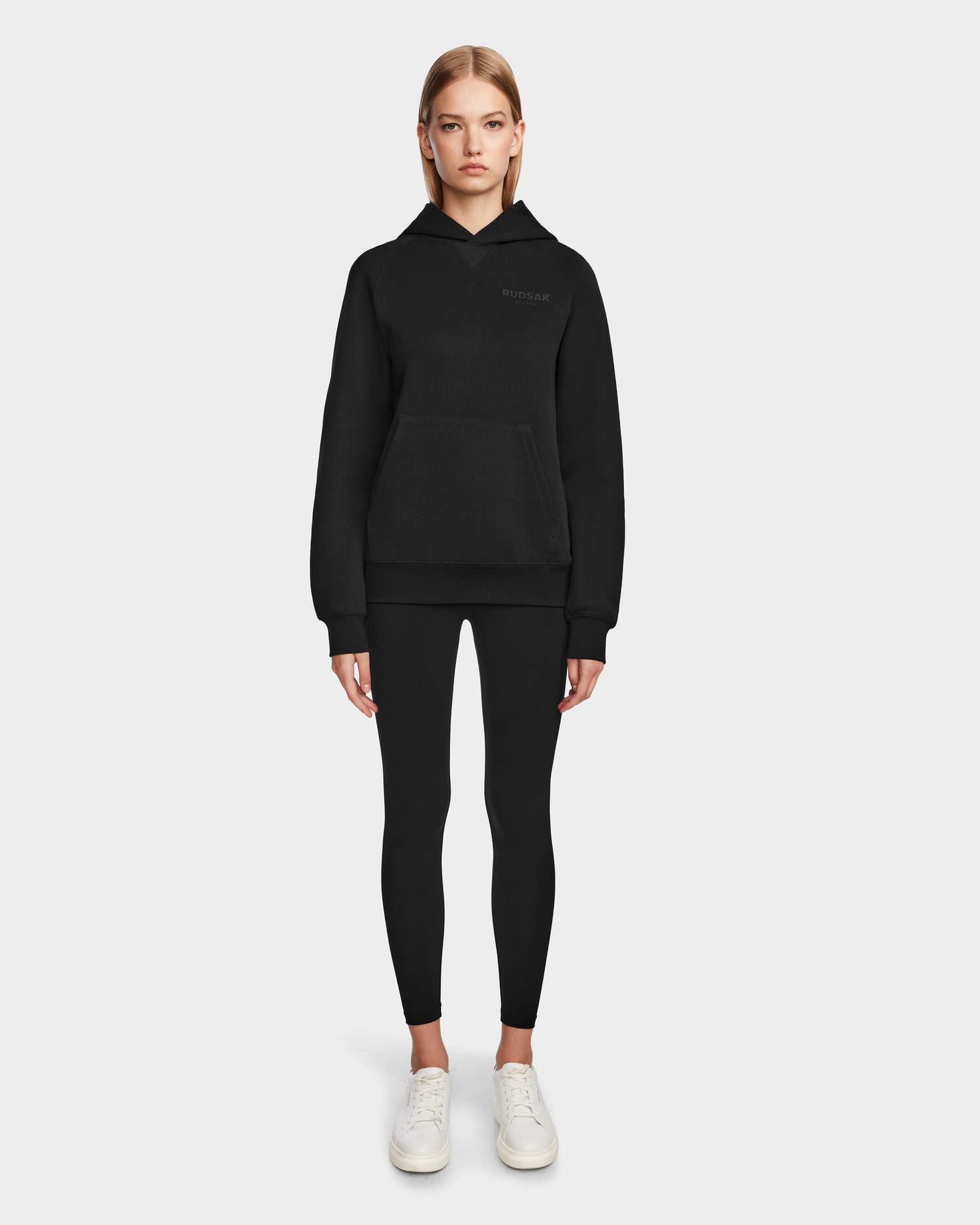 A woman faces forward against a plain light gray background, wearing the Rudsak BARRY - 9524011 Black W oversized hoodie, paired with black leggings and white sneakers.