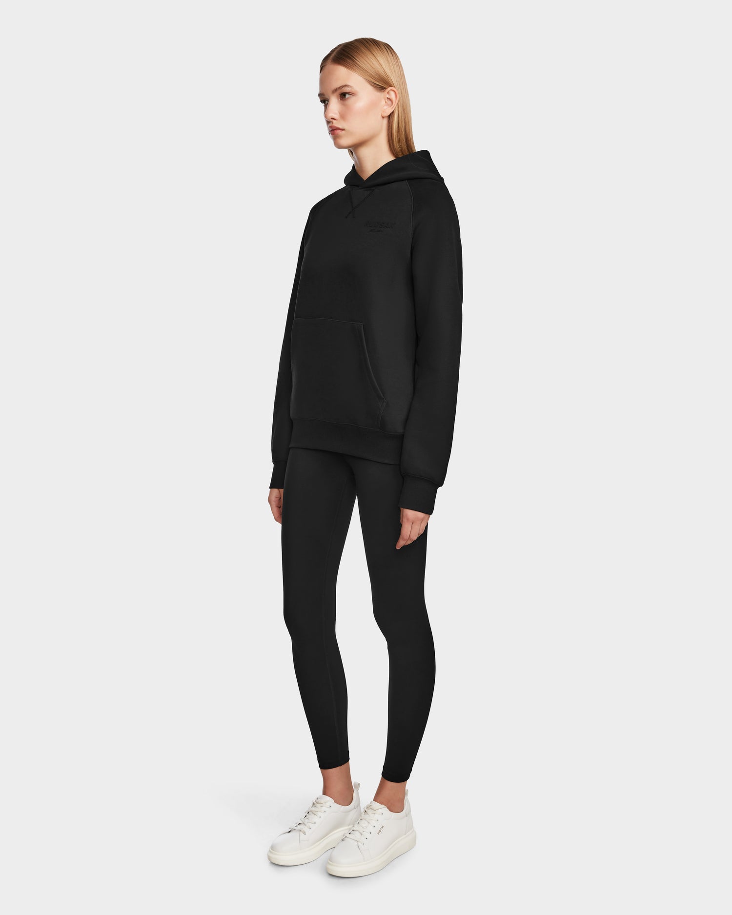 A woman with straight, shoulder-length blonde hair and a neutral expression stands against a plain background wearing the Rudsak BARRY - 9524011 Black W hoodie, black leggings, and white sneakers.