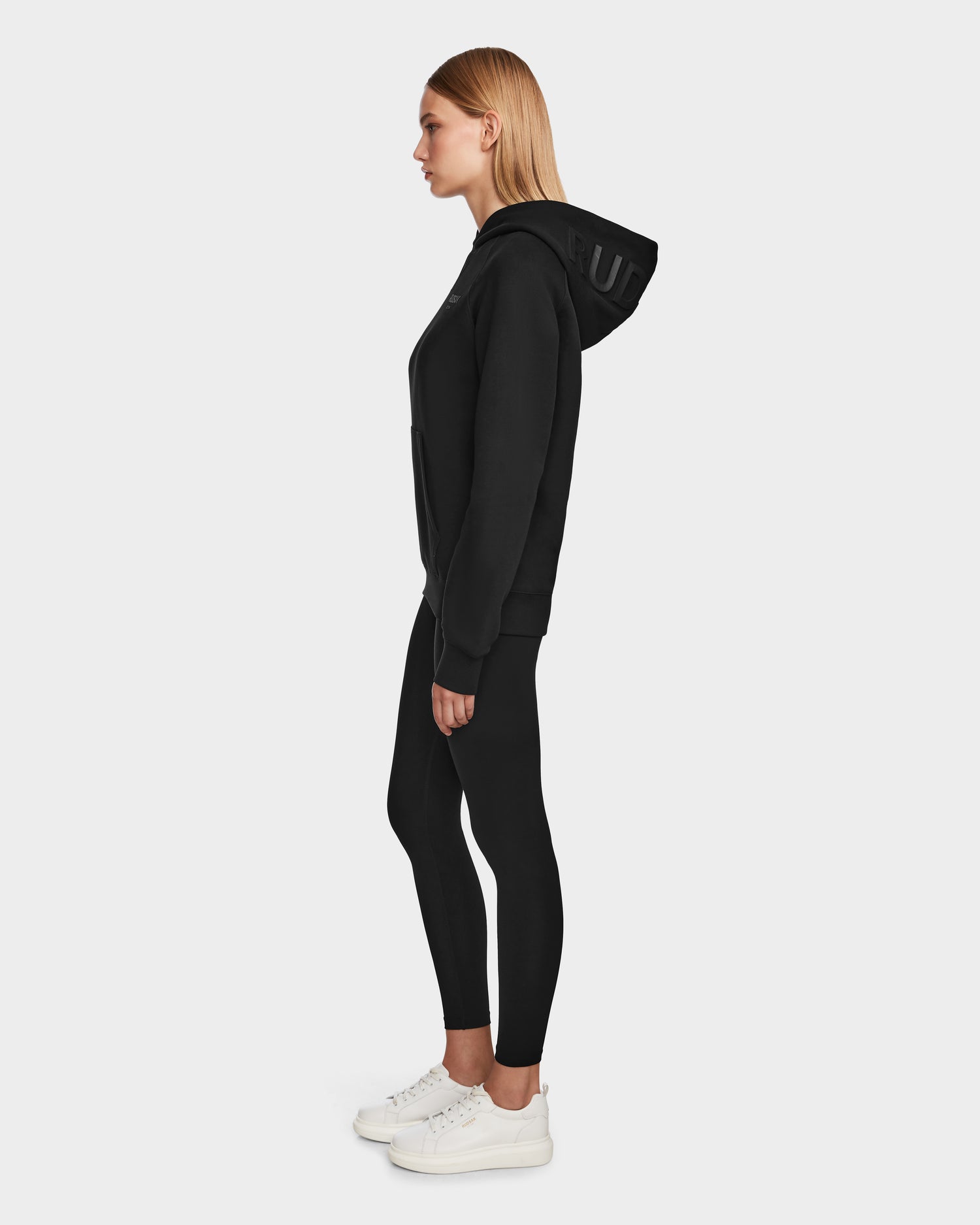 A woman stands in profile wearing a Rudsak BARRY - 9524011 Black W oversized hoodie, paired with black leggings and white sneakers, against a plain light background.