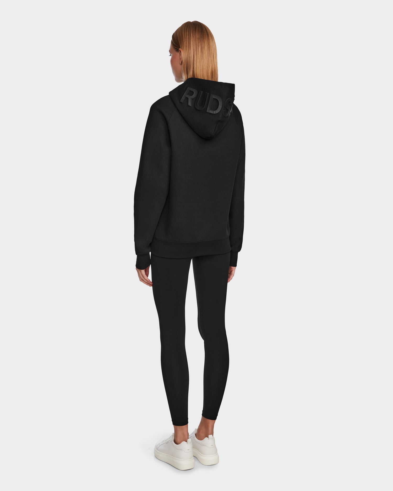 A woman faces away in the Rudsak BARRY - 9524011 Black W hoodie with “RUDS” on the hood, paired with black leggings and white sneakers against a plain light background.