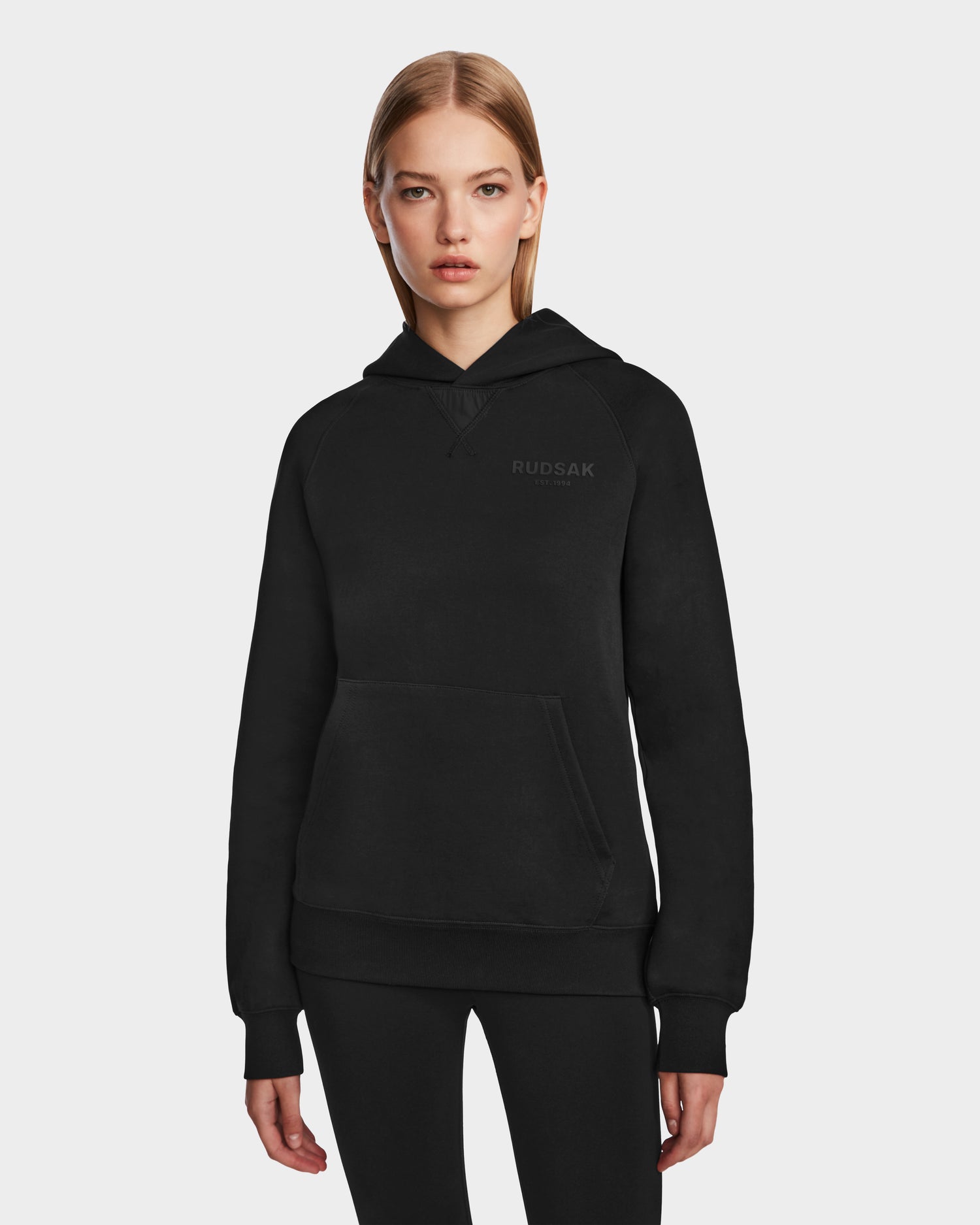 A woman with straight blonde hair wears the Rudsak BARRY - 9524011 Black W oversized hoodie featuring a front pocket and “RUDSAK” logo, paired with black pants, standing against a plain white background.