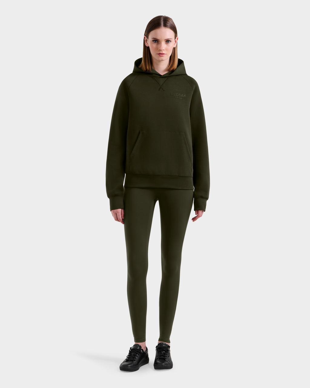 A woman faces forward wearing the Rudsak BARRY - 9524011 Dark Olive W hoodie and matching leggings with black sneakers, set against a plain light gray background.