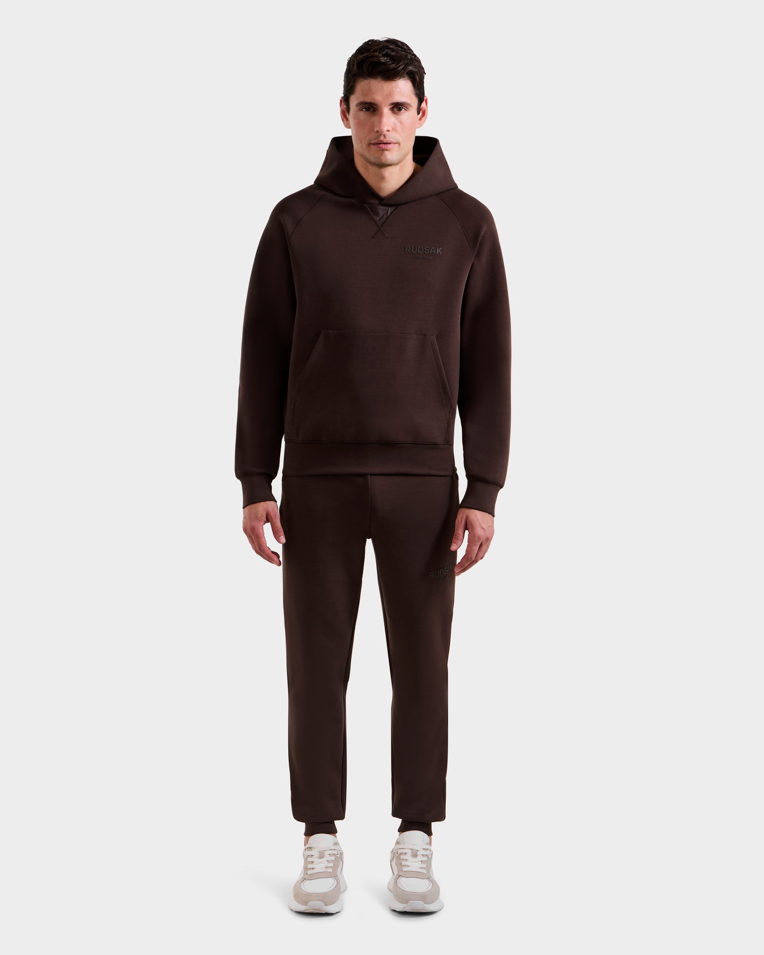 A man stands facing forward wearing the Rudsak BARRY - 9524011 Espresso M hoodie and matching jogger pants with white sneakers, set against a plain white background.