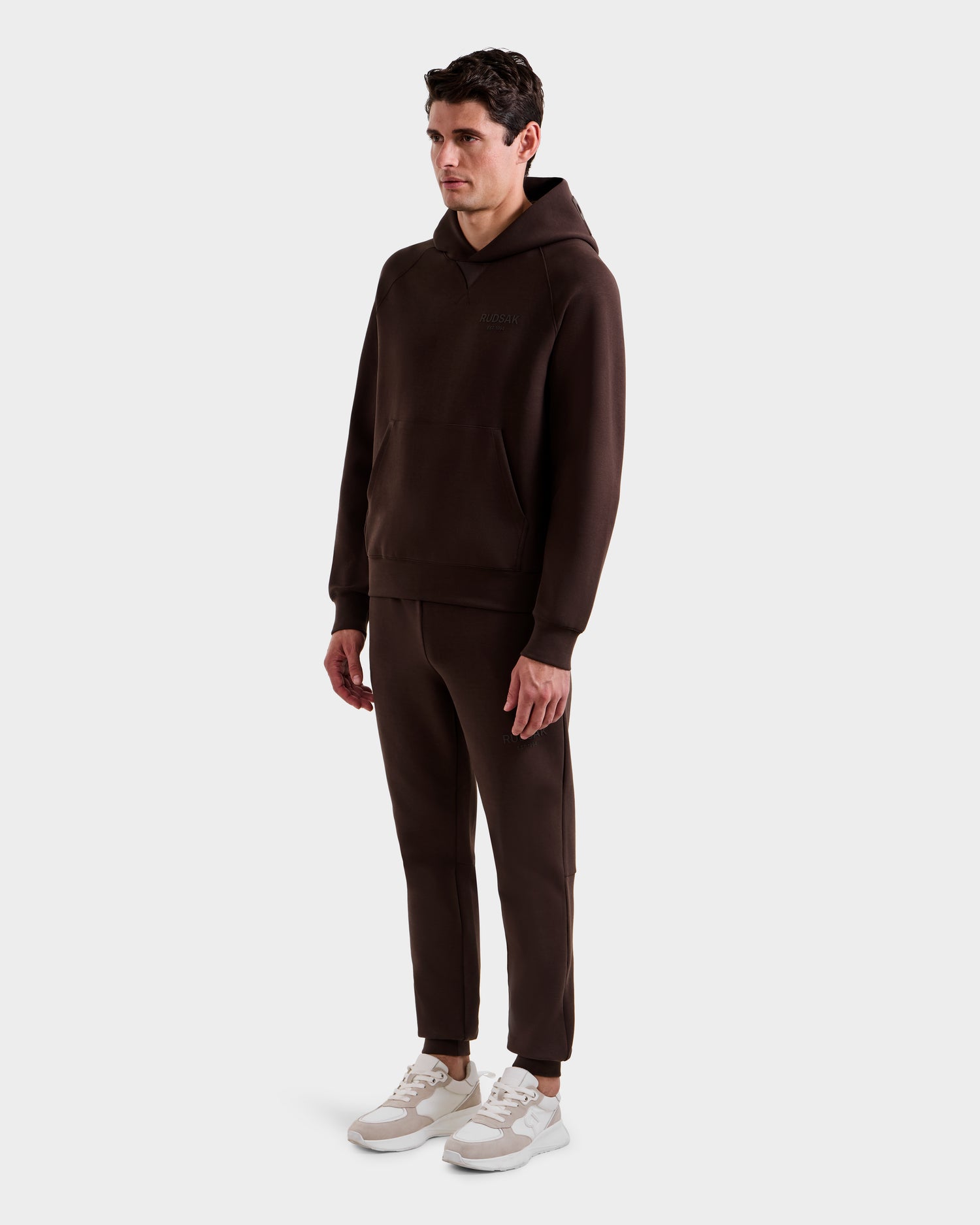A man stands against a plain light gray background wearing the Rudsak BARRY - 9524011 Espresso M hoodie and matching jogger pants, paired with white sneakers.