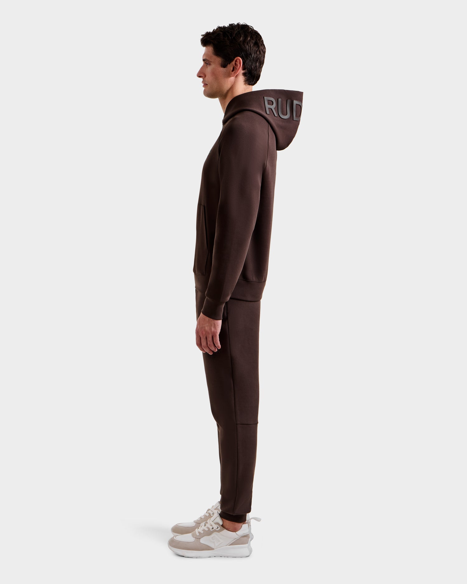 A man stands in profile wearing the Rudsak BARRY - 9524011 Espresso M hoodie and matching joggers, with white sneakers, set against a plain light gray background.