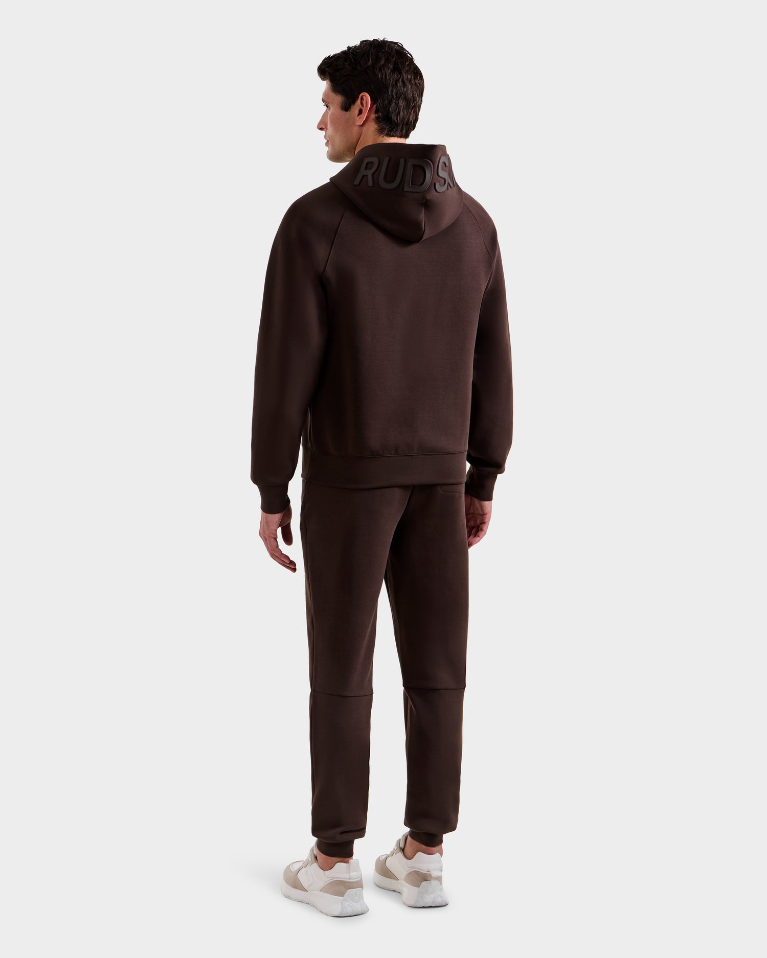 A man faces away wearing the Rudsak BARRY - 9524011 Espresso M hoodie and matching joggers, hood up to show "RUDDS" boldly on the back. He pairs the oversized unisex set with white sneakers, standing against a plain white background.
