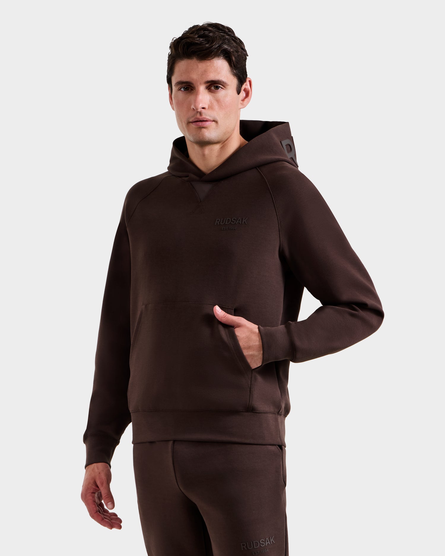 A man models the Rudsak BARRY - 9524011 Espresso M unisex hoodie and sweatpants set, featuring "RUDSAK" on the chest, standing against a plain white background with one hand in his pocket.