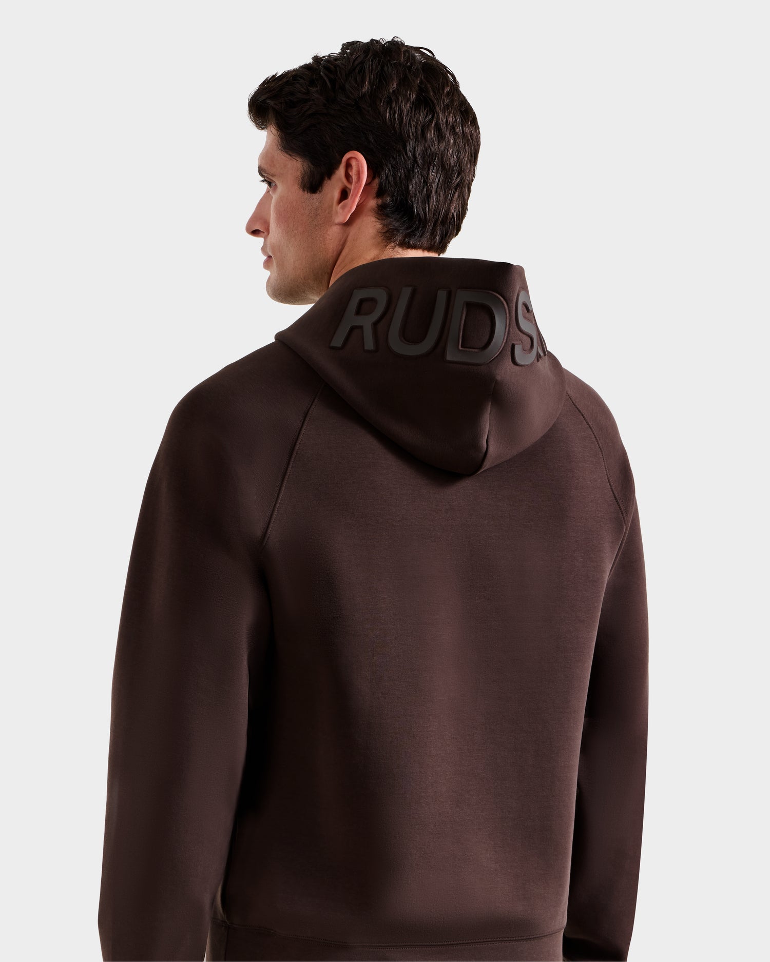 A man with short dark hair is seen from the back wearing the Rudsak BARRY - 9524011 Espresso M unisex hoodie, featuring "RUDSAK" embossed on the hood, against a plain light background.