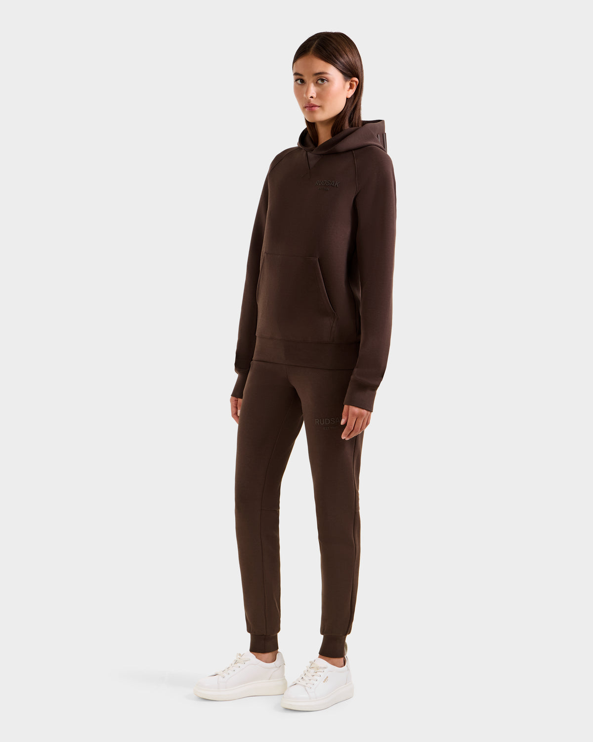A woman faces forward wearing the Rudsak BARRY - 9524011 Espresso W oversized hoodie and jogger set with white sneakers, against a plain light background.