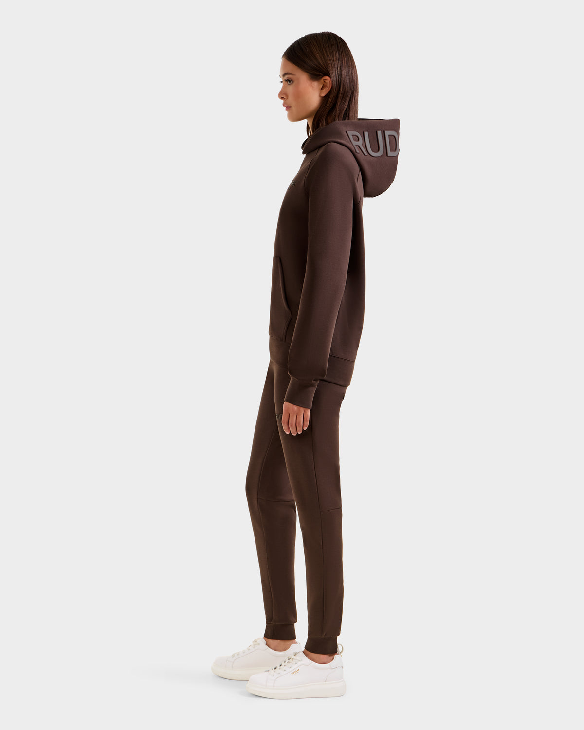 A woman stands in profile wearing the Rudsak BARRY - 9524011 Espresso W oversized hoodie and joggers set with white sneakers. The hoodie has bold lettering on the hood, and the background is plain white.
