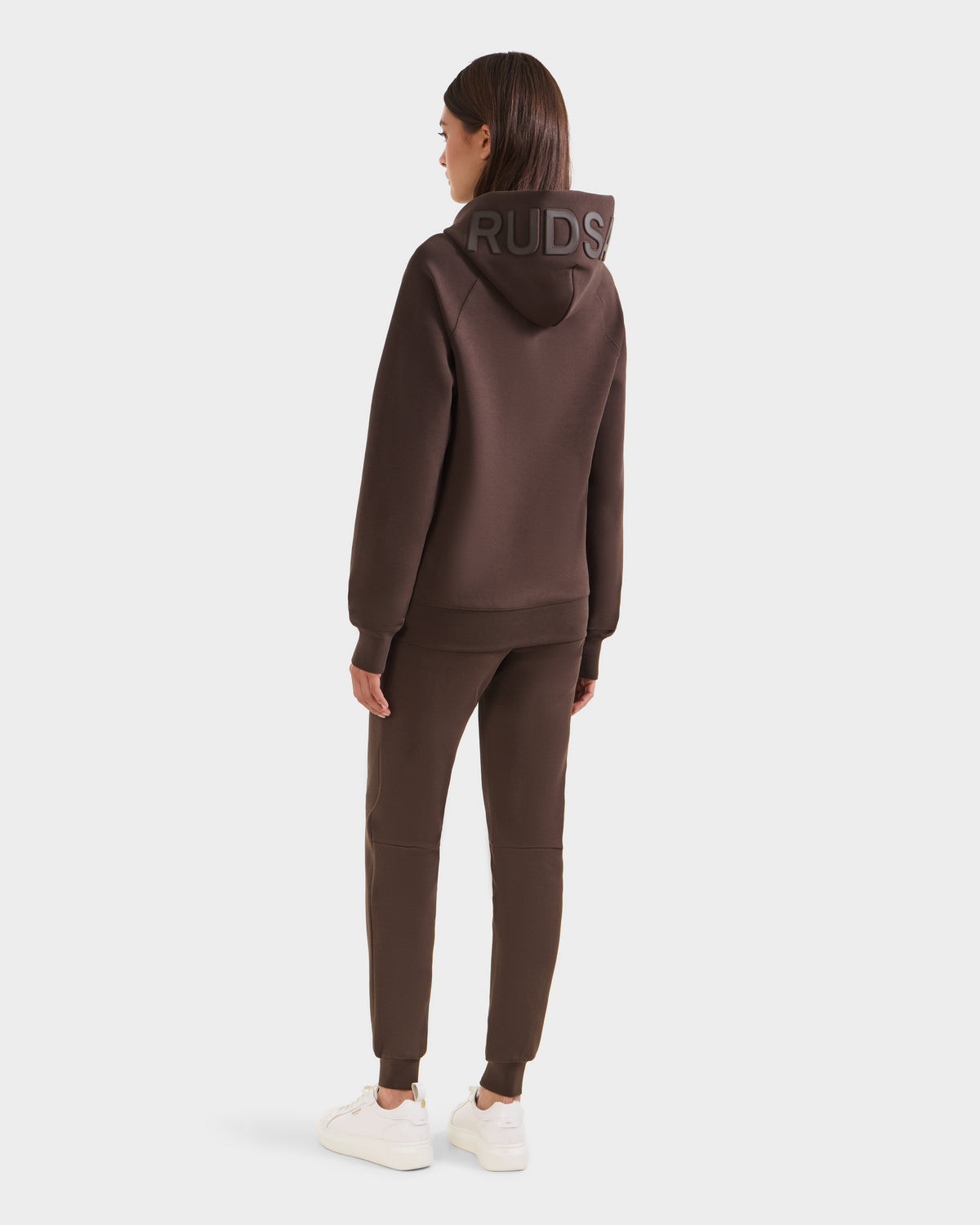 A woman faces away, wearing the Rudsak BARRY - 9524011 Espresso W oversized hoodie and jogger pants with white sneakers. The hoodie’s hood displays large block letters against a plain white background.