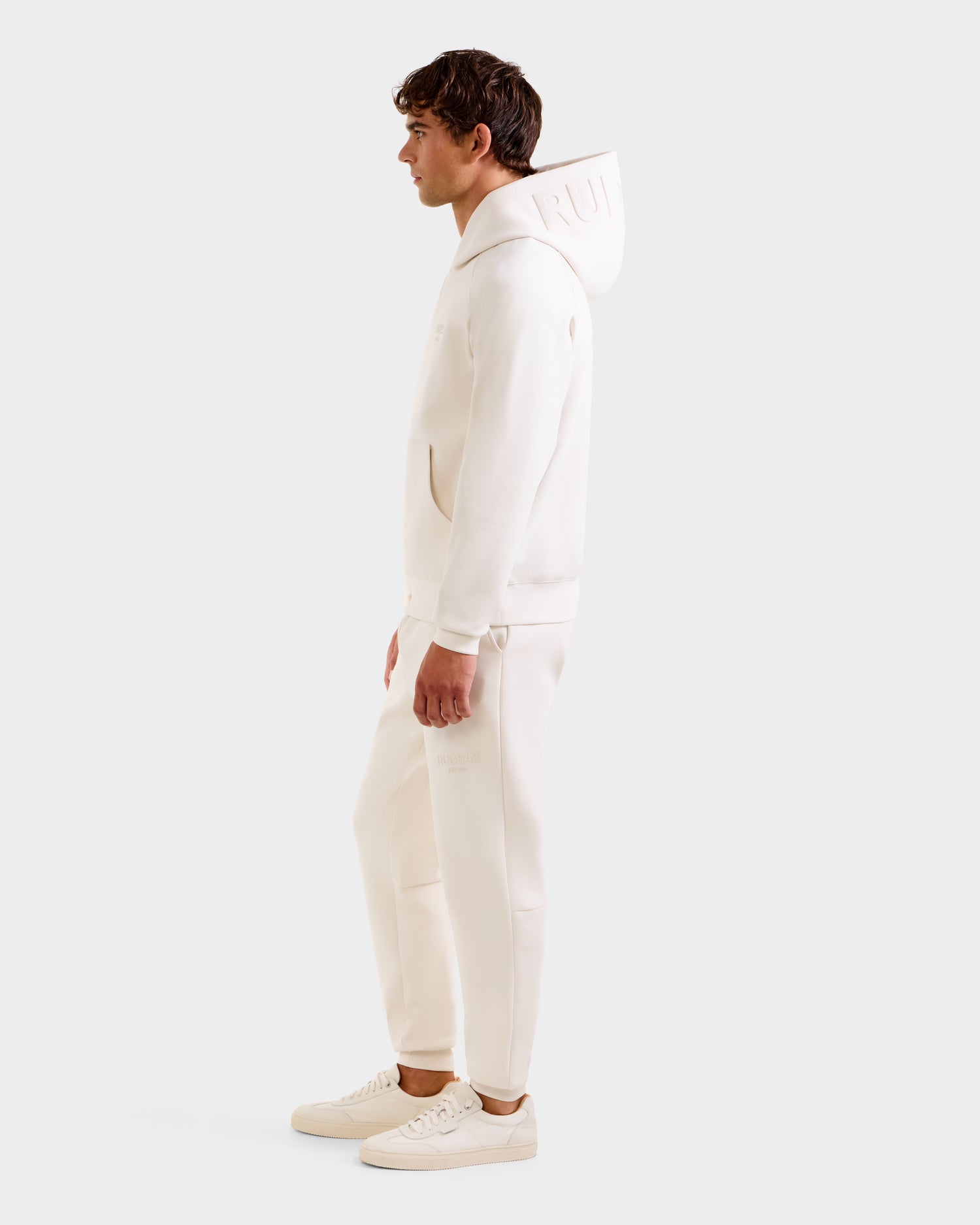 A man stands in profile against a plain white background, wearing the Rudsak BARRY - 9524011 Vanilla M oversized unisex hoodie, paired with matching sweatpants and sneakers for an all-white look.