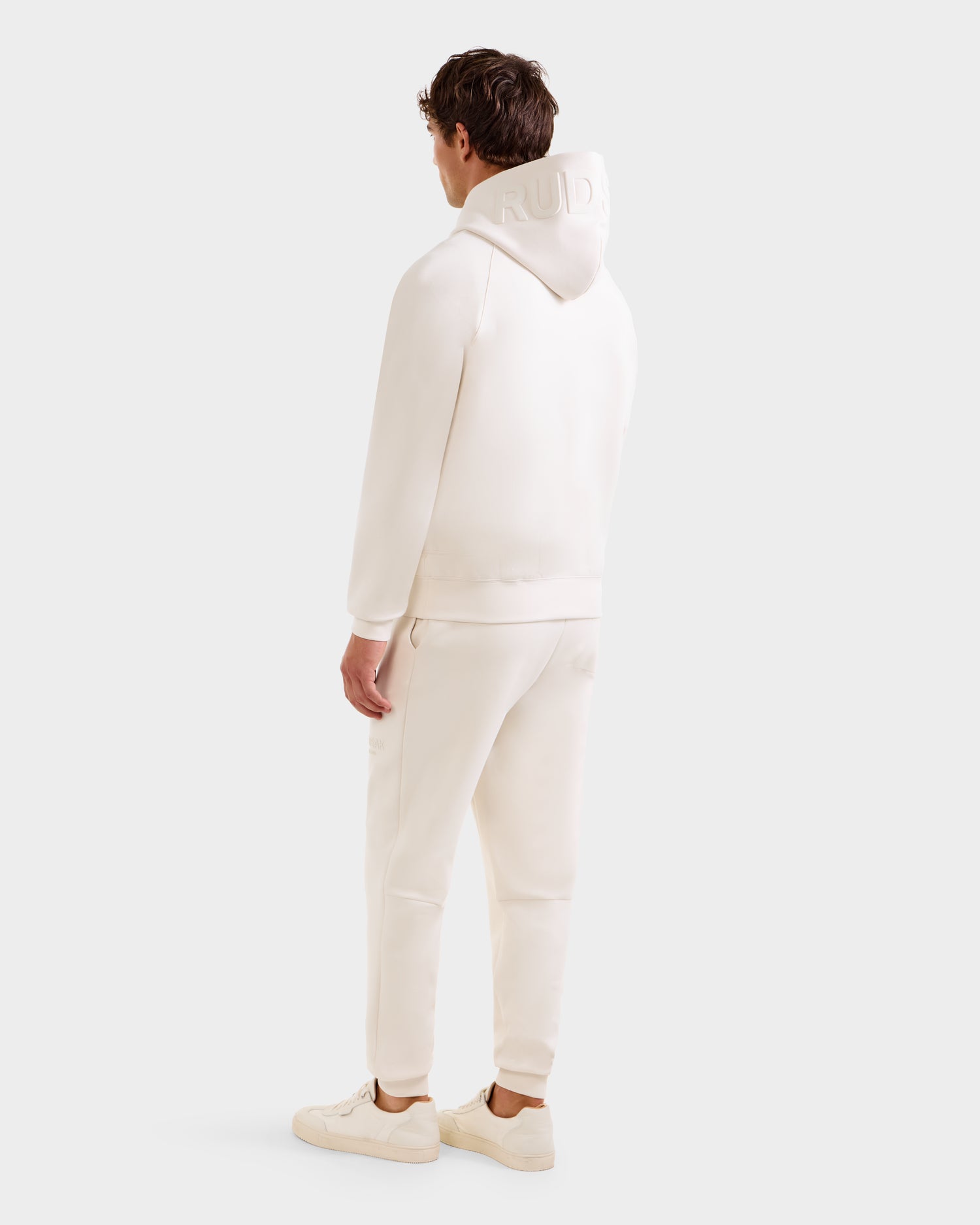 A person stands facing away, wearing the oversized Rudsak BARRY - 9524011 Vanilla M hoodie with the hood up, matching jogger pants, and off-white sneakers against a plain, light background.