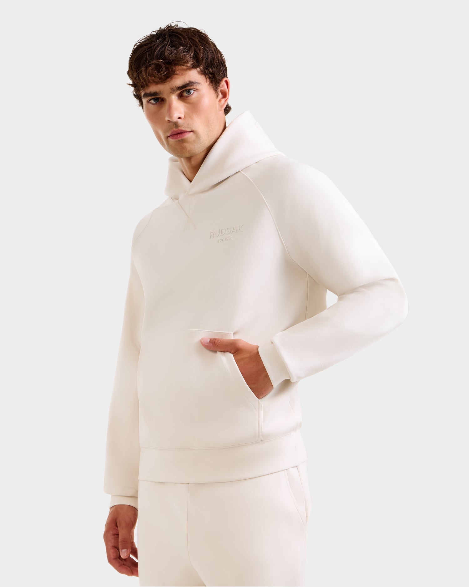 A young man with short brown hair wears the Rudsak BARRY - 9524011 Vanilla M hoodie and matching pants, standing against a plain light background with one hand in his pocket, looking seriously at the camera.