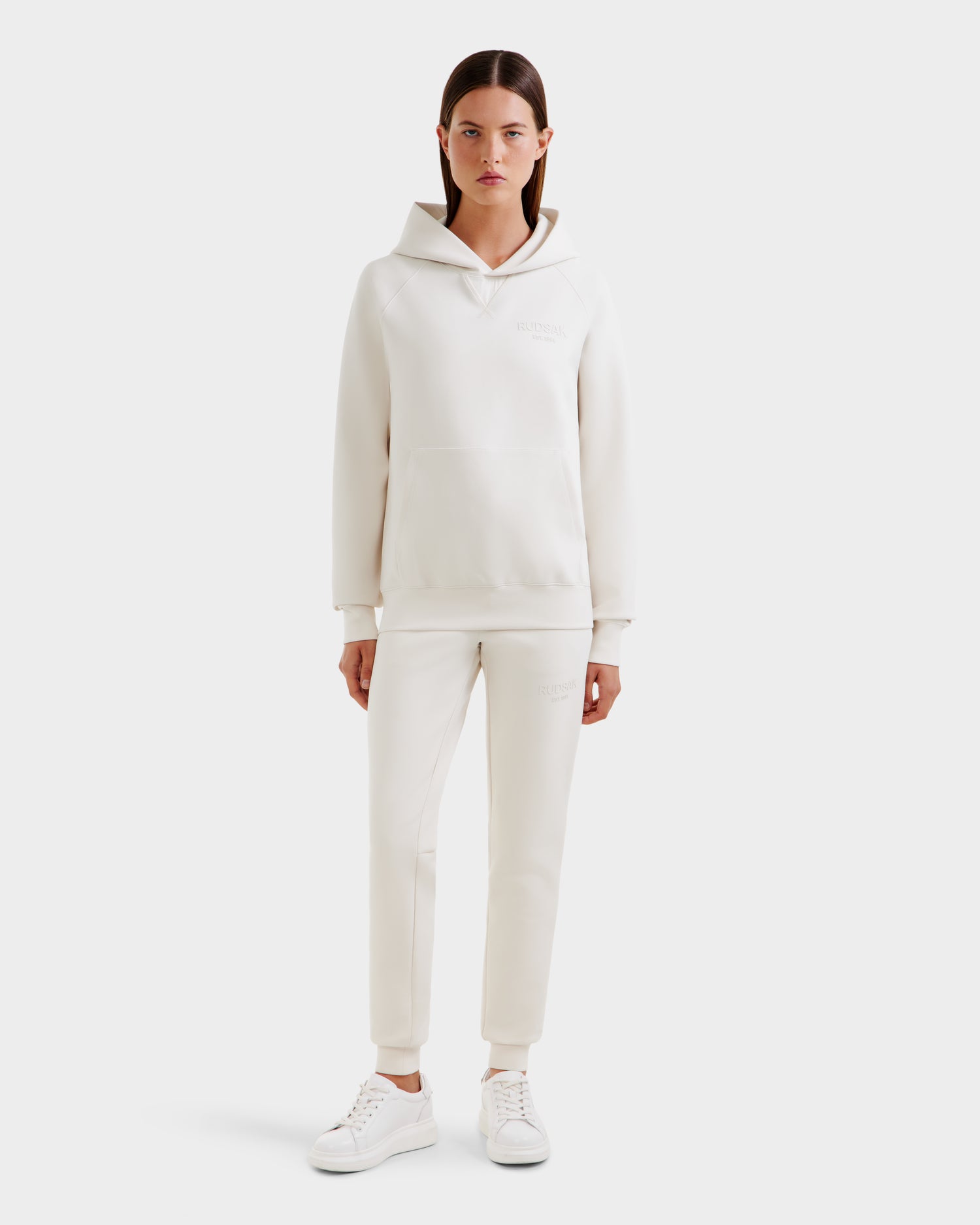 A woman stands facing forward, wearing the Rudsak BARRY - 9524011 Vanilla W oversized white hoodie with matching jogger pants and white sneakers against a plain light-colored background.