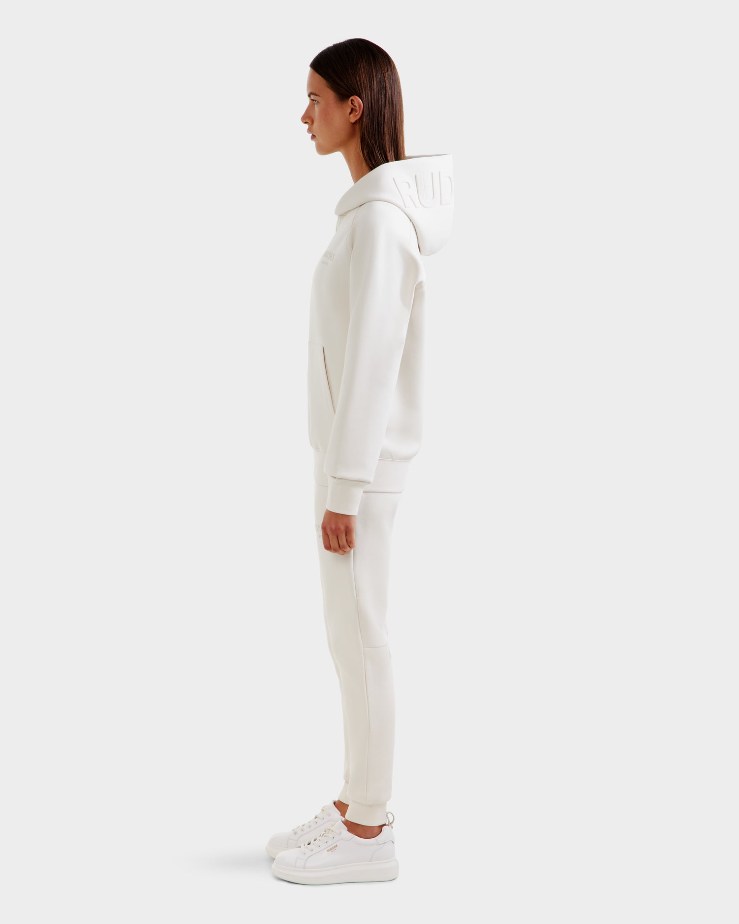 A woman stands in profile against a plain background, wearing the Rudsak BARRY - 9524011 Vanilla W oversized white hoodie and matching pants. Her straight dark brown hair is down, and her hands rest by her sides.