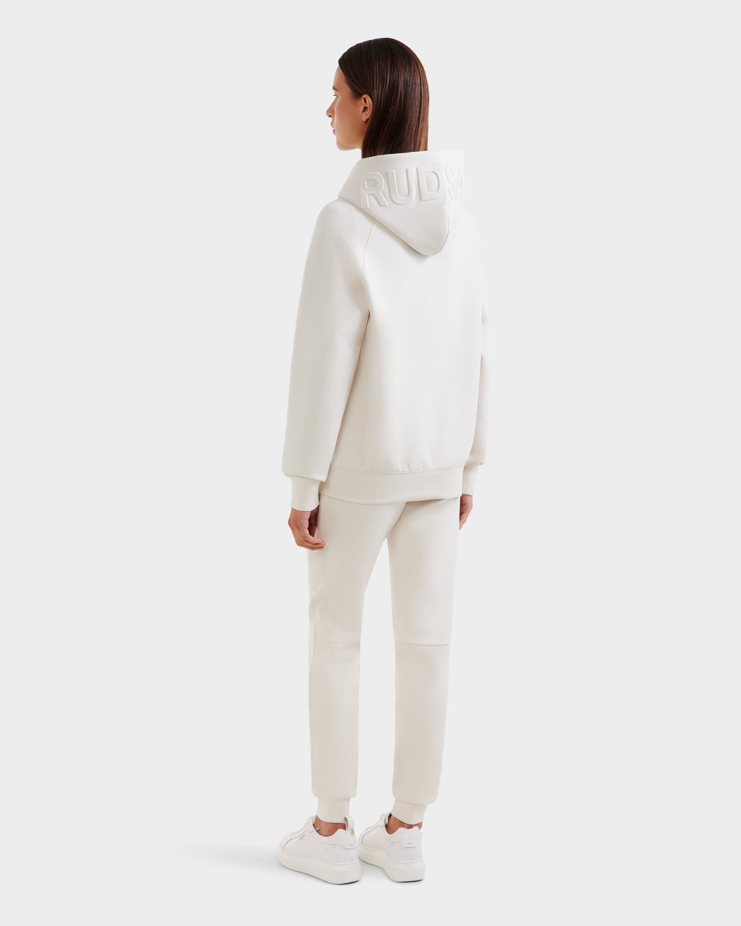 A woman faces away, wearing the Rudsak BARRY - 9524011 Vanilla W hoodie with embossed hood text, paired with matching white joggers and sneakers against a plain, light background.