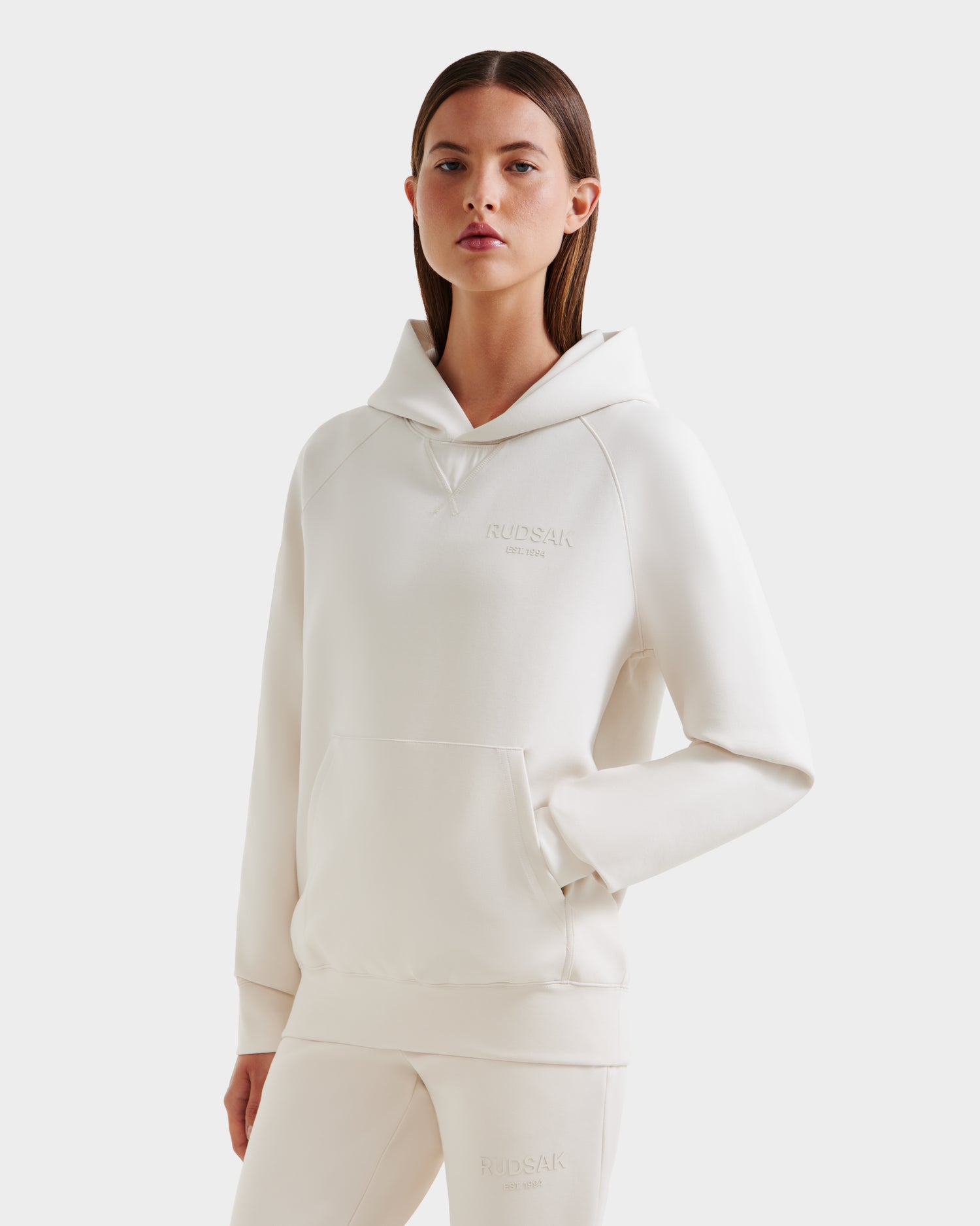 A woman stands with one hand in her pocket, wearing the oversized Rudsak BARRY - 9524011 Vanilla W unisex hoodie and matching pants. The hoodie features a front pocket and subtle chest logo, set against a plain white background.
